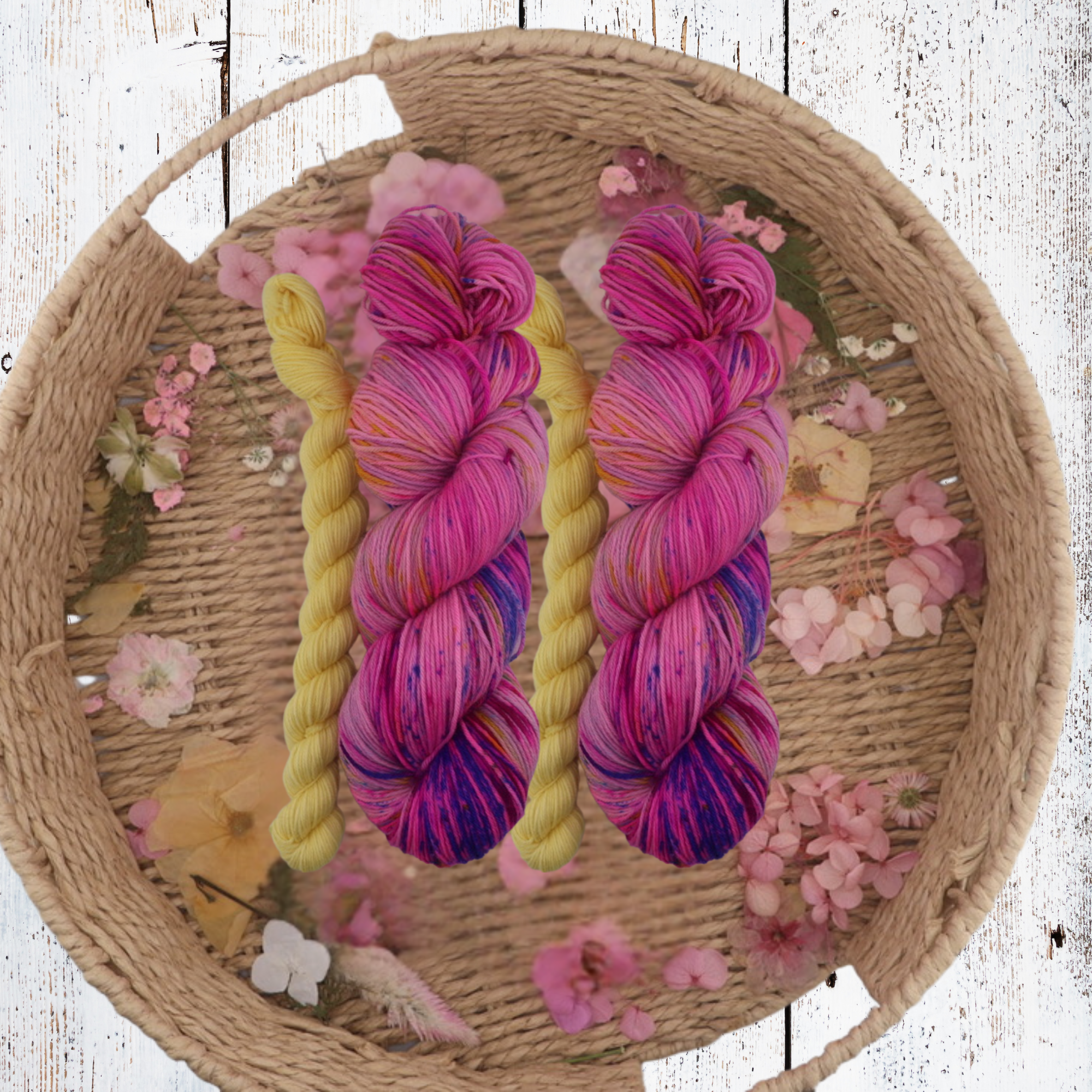 Hand‑dyed yarn in pink, purple, magenta and blue shades arranged in a basket with dried petals, showing tonal depth and soft halo on a rustic white surface.