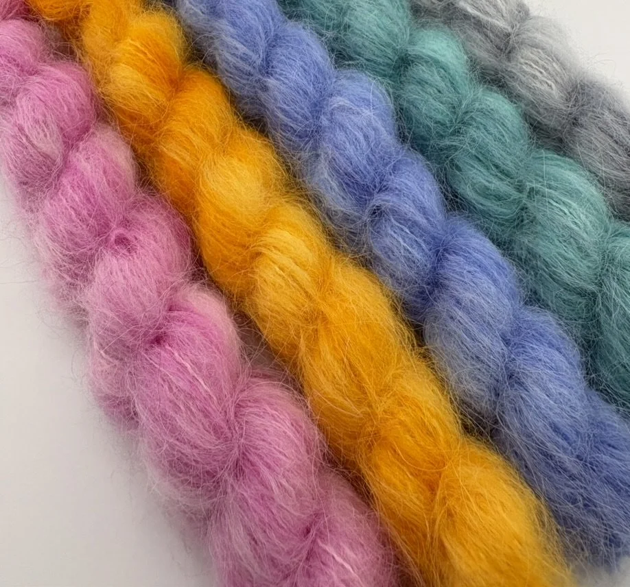 Close-up of four colorful, fluffy yarns in pink, yellow, blue, and teal, arranged in parallel.