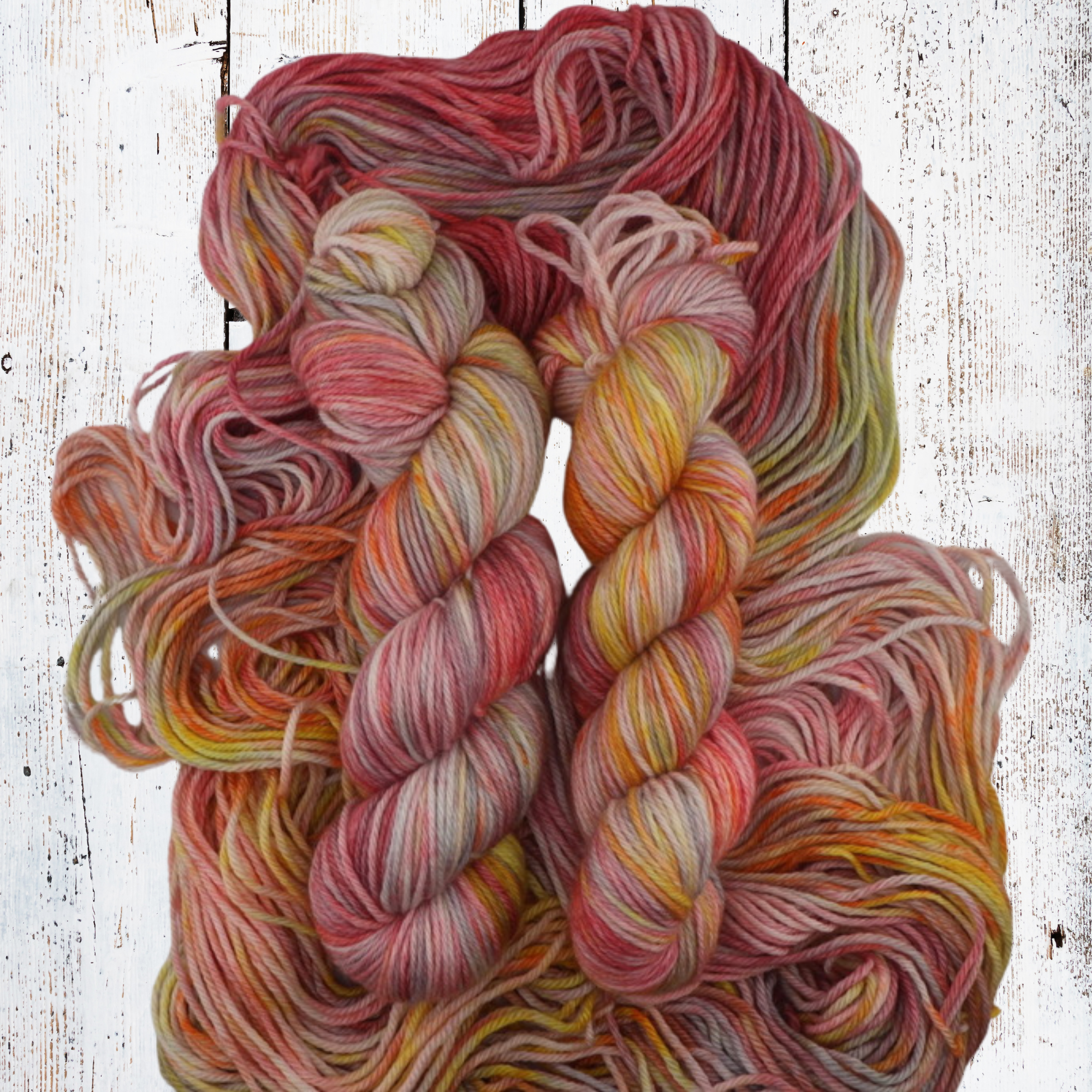 Multicoloured yarn skeins in warm pink, orange, yellow and purple shades displayed in a woven basket with scattered flowers on a rustic white wooden surface