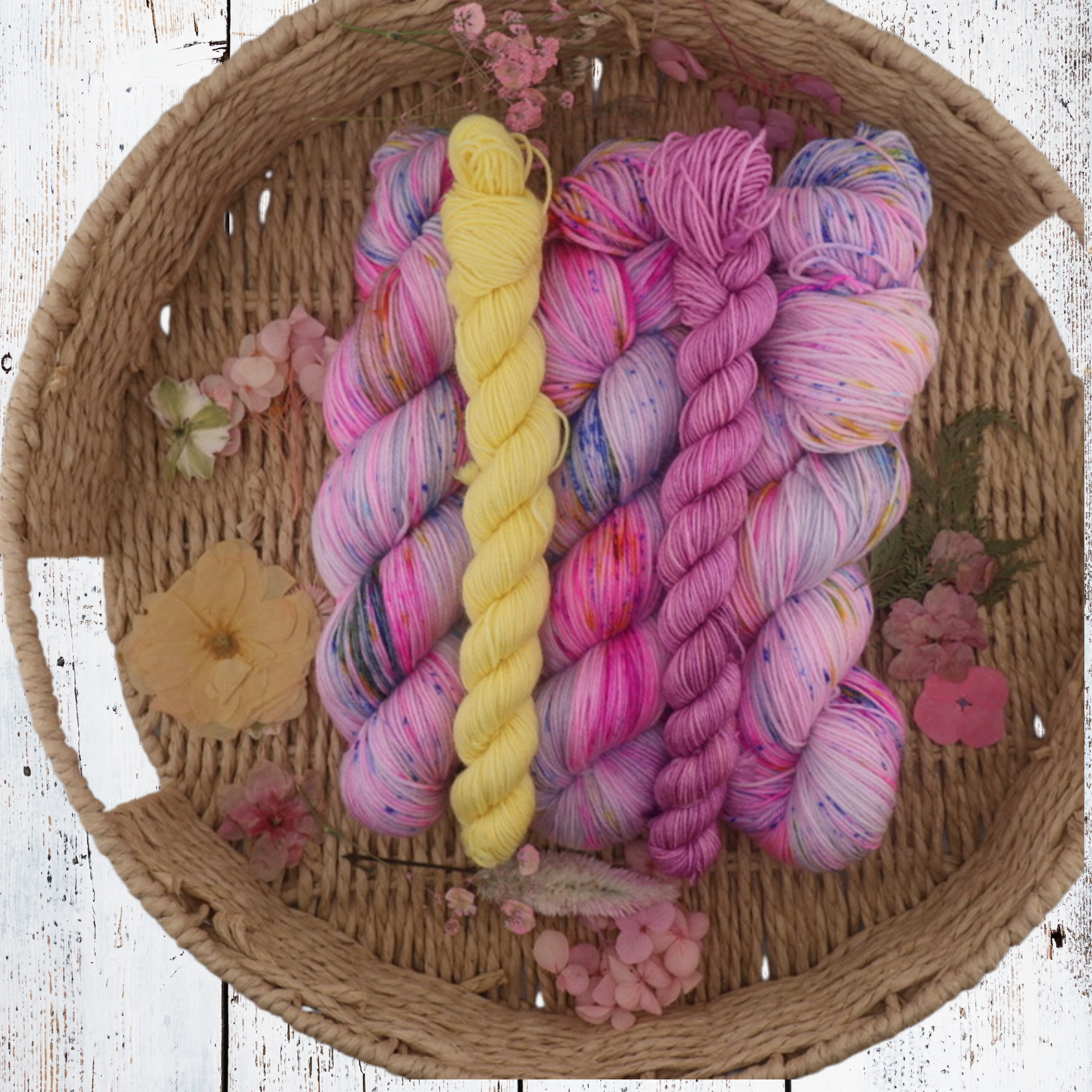 Bright pink, purple, blue and yellow hand‑dyed yarn skeins styled together in a wicker basket with scattered petals, showing rich colour variation and fibre softness.