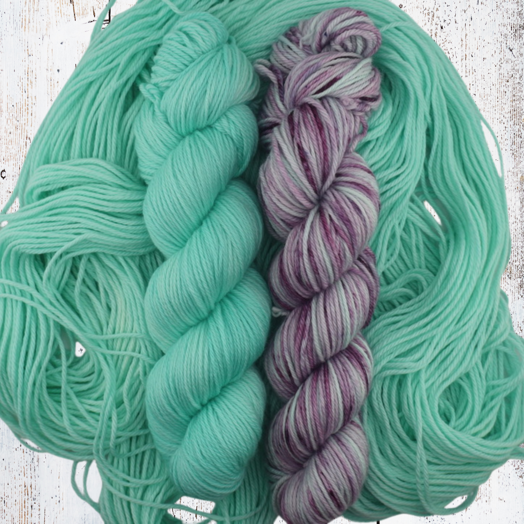 Purple, grey and mint‑green hand‑dyed yarn skeins arranged on a white wooden surface with loose fibres, highlighting tonal variation across the colourway.