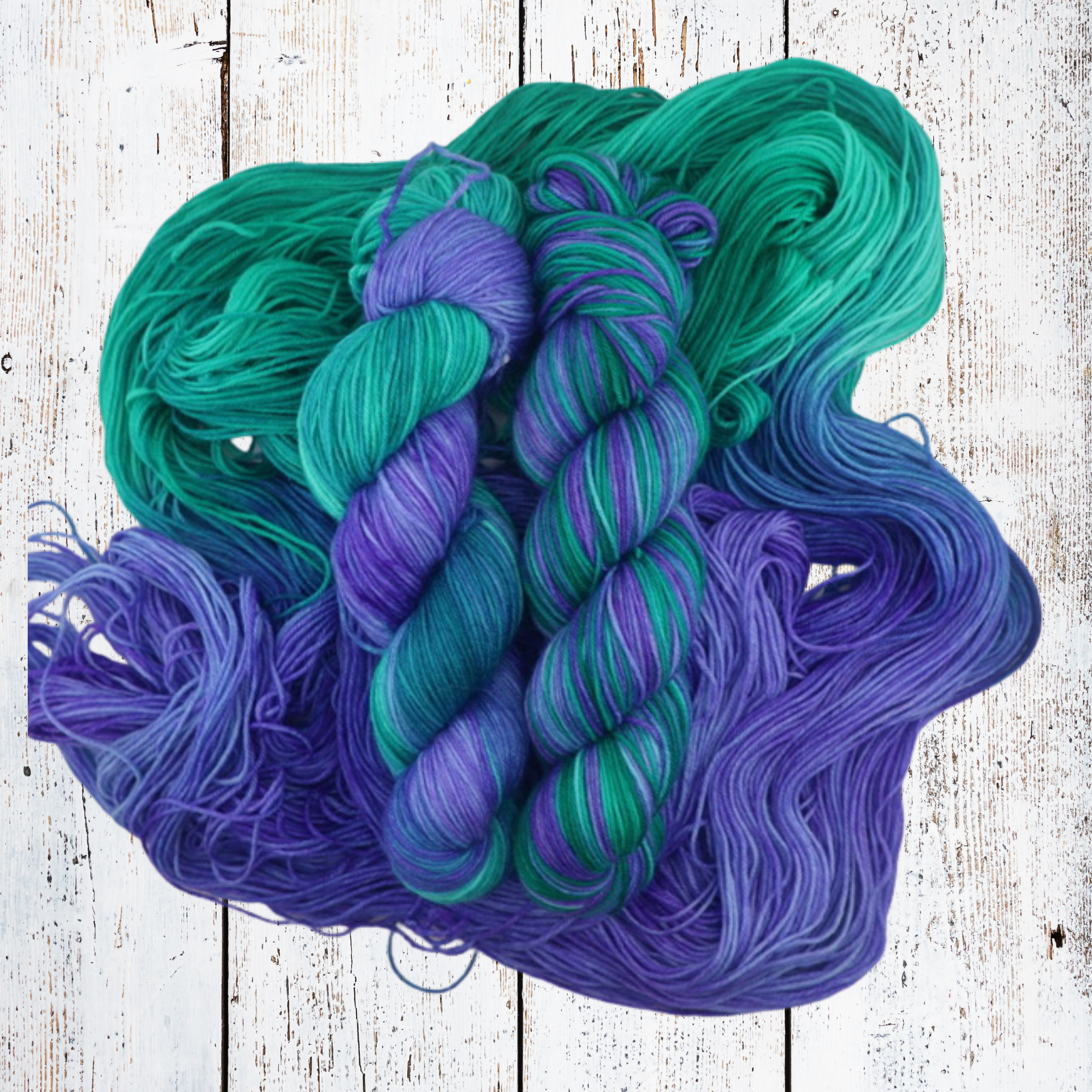 Loose hand‑dyed yarn and skeins in teal, blue and purple shades arranged on a white wooden background to showcase tonal variation and fibre softness.