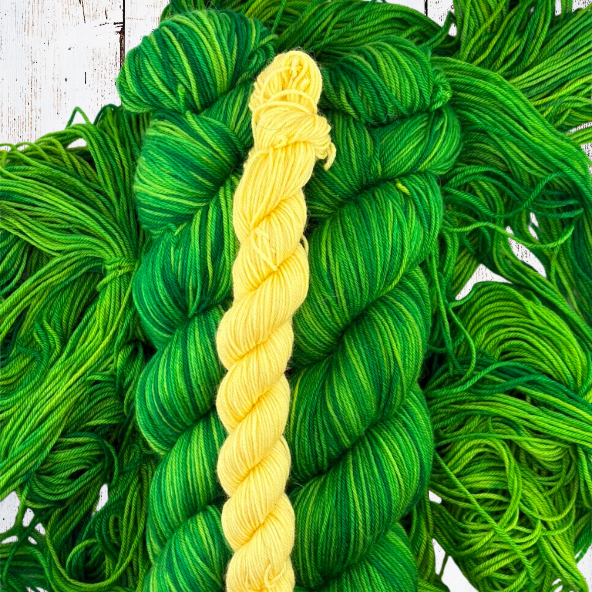 Hand‑dyed 75/25% Merino/Nylon yarn in green tonal shades with a yellow skein in the centre, arranged on a flat surface to show texture and colour contrast.