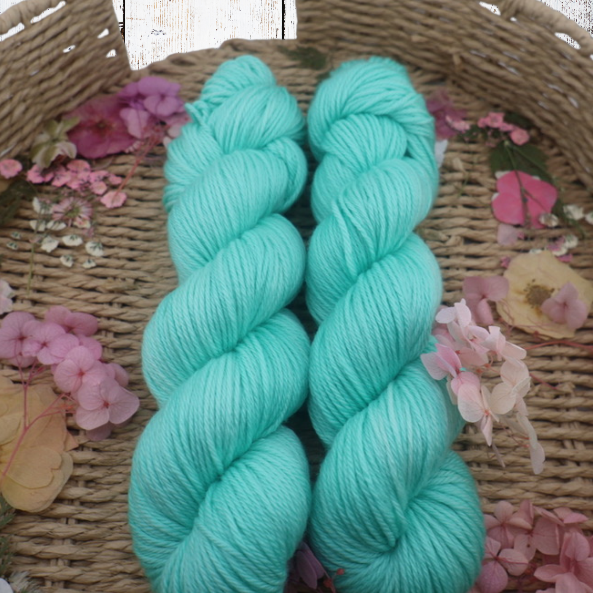 Close‑up of two saturated turquoise hand‑dyed yarn skeins arranged in a woven basket with scattered dried petals.
