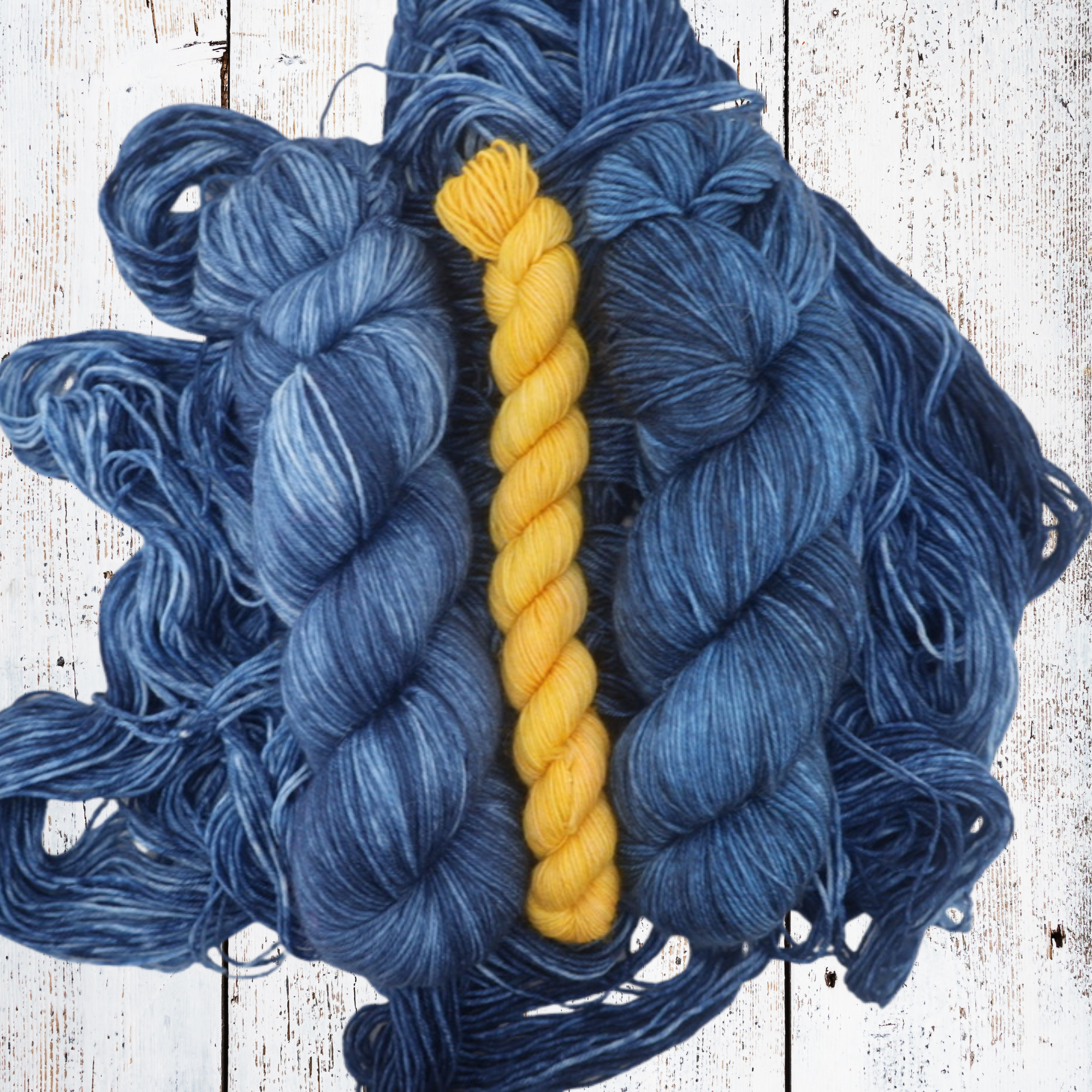 Multiple skeins of hand‑dyed blue yarn spread out on a white wooden background with a single yellow skein placed on top, showcasing colour contrast and yarn texture.