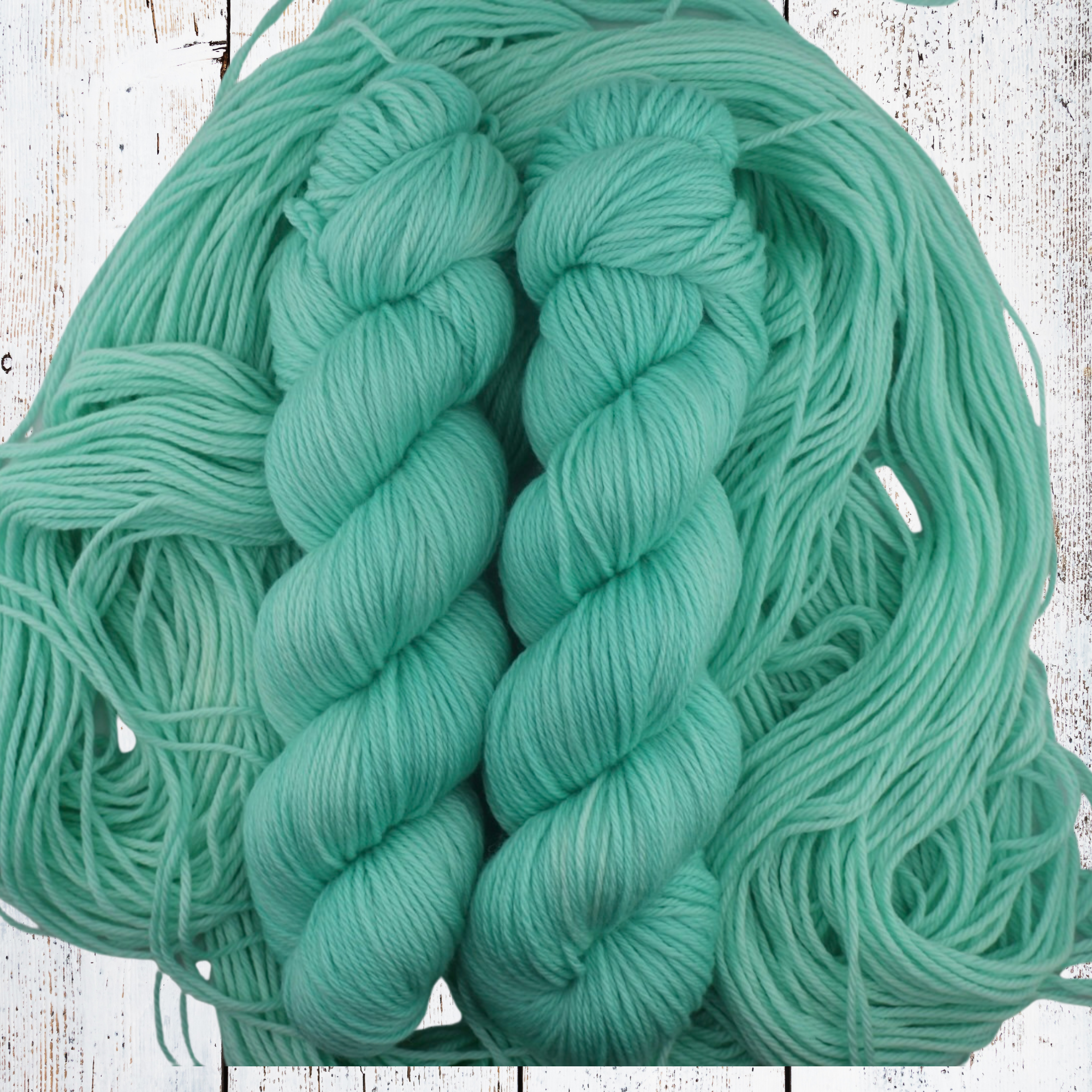 Three turquoise yarn skeins displayed on a white wooden surface, surrounded by loose strands of matching fibre.