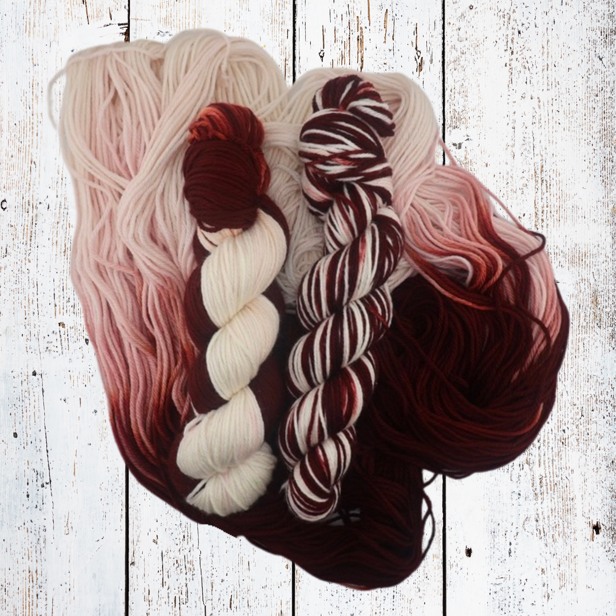 Multiple hand‑dyed yarn skeins in burgundy, cream and pink tones laid out on a white wooden surface, highlighting tonal variation across the colourway