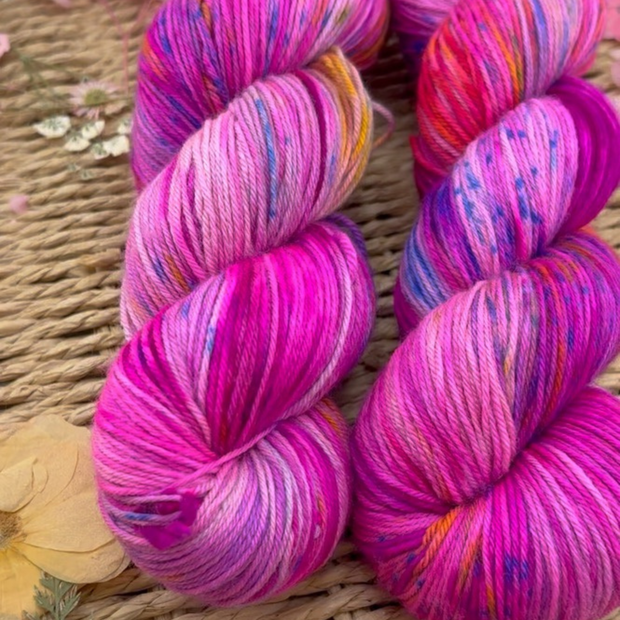Multicoloured pink, purple and magenta yarn skeins with a contrasting yellow skein in a woven basket, styled with scattered petals to showcase texture and vibrant colour blends.