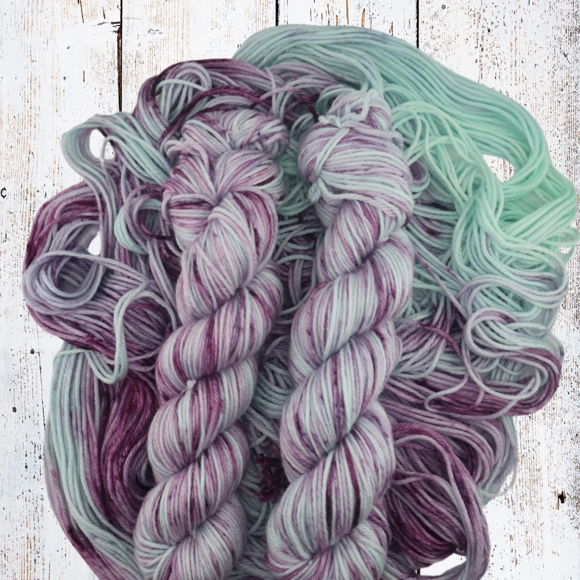 Multiple hand‑dyed yarn skeins in purple, grey and mint tones laid out on a white wooden background, showing soft fibre texture and colour transitions.