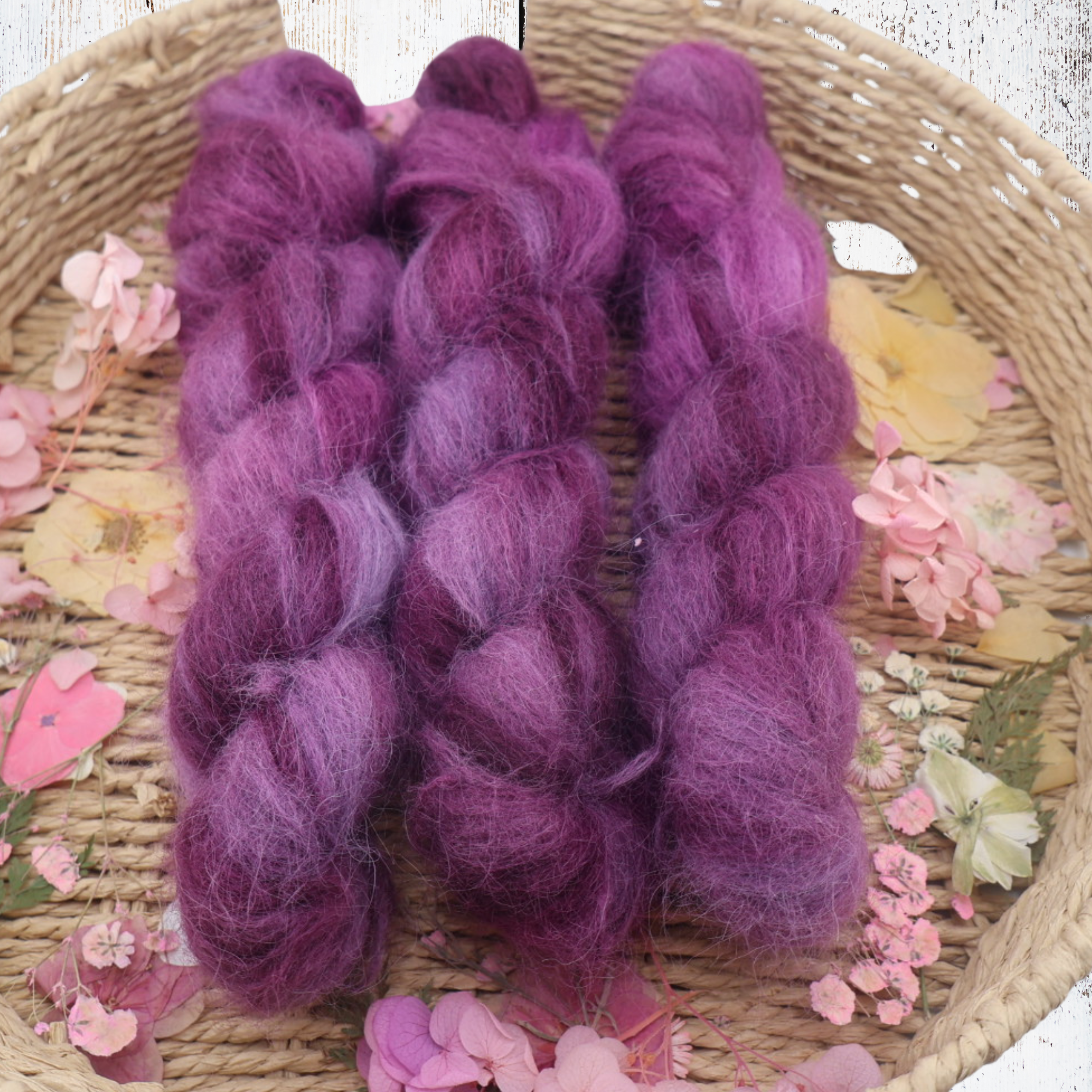 Close‑up of fluffy purple alpaca yarn skeins with rich violet and lavender tones, displayed in a rustic basket with scattered dried petals for craft photography.