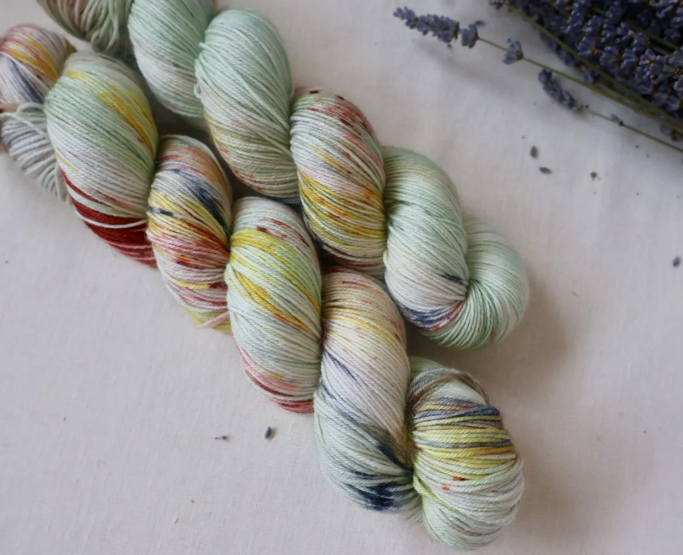 A skein of multicolored yarn with specks of red, yellow, green, blue, and black on a white background. A bunch of dried lavender flowers is visible in the top right corner.