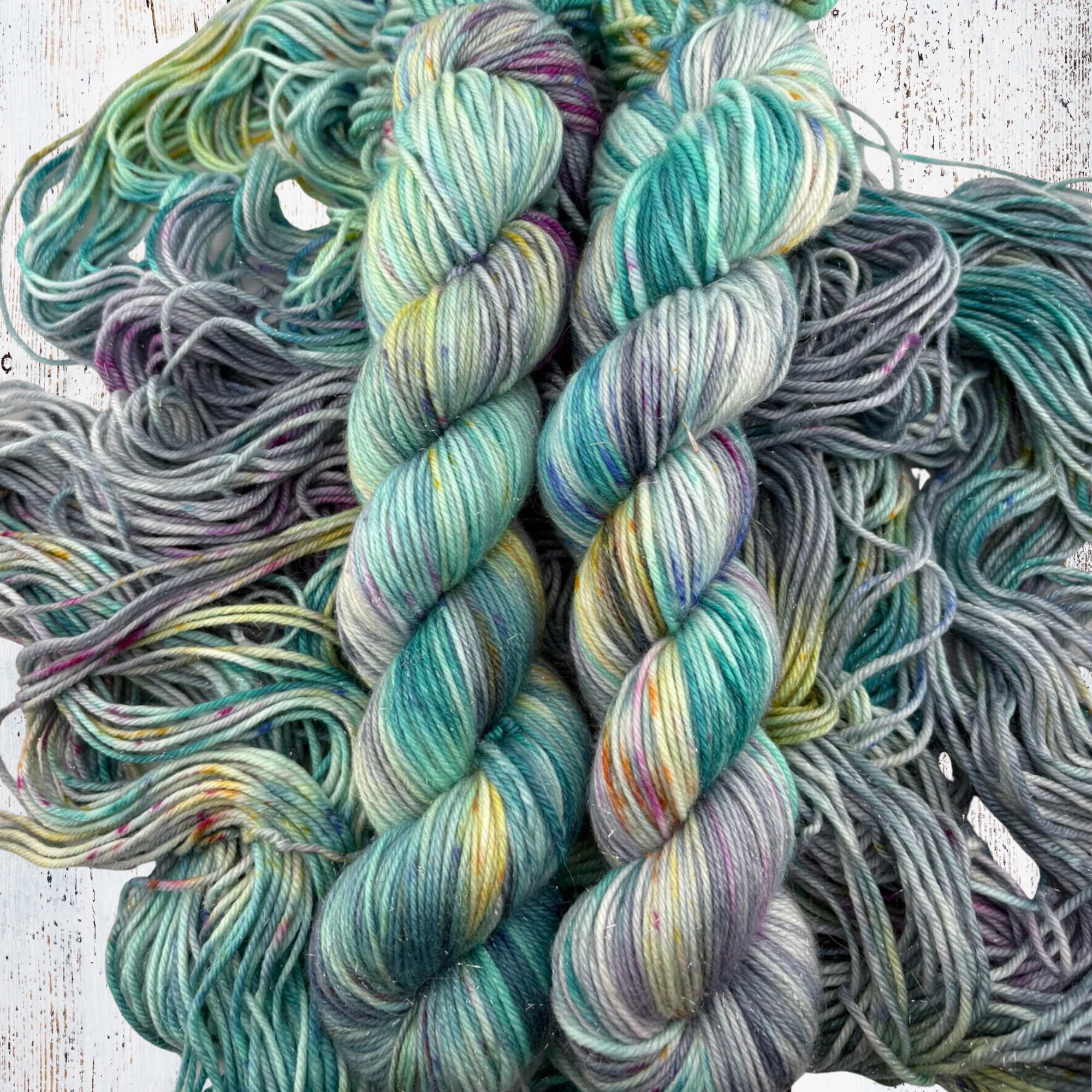 Close‑up of hand‑dyed yarn in turquoise, green, and purple shades on a textured surface, highlighting tonal depth