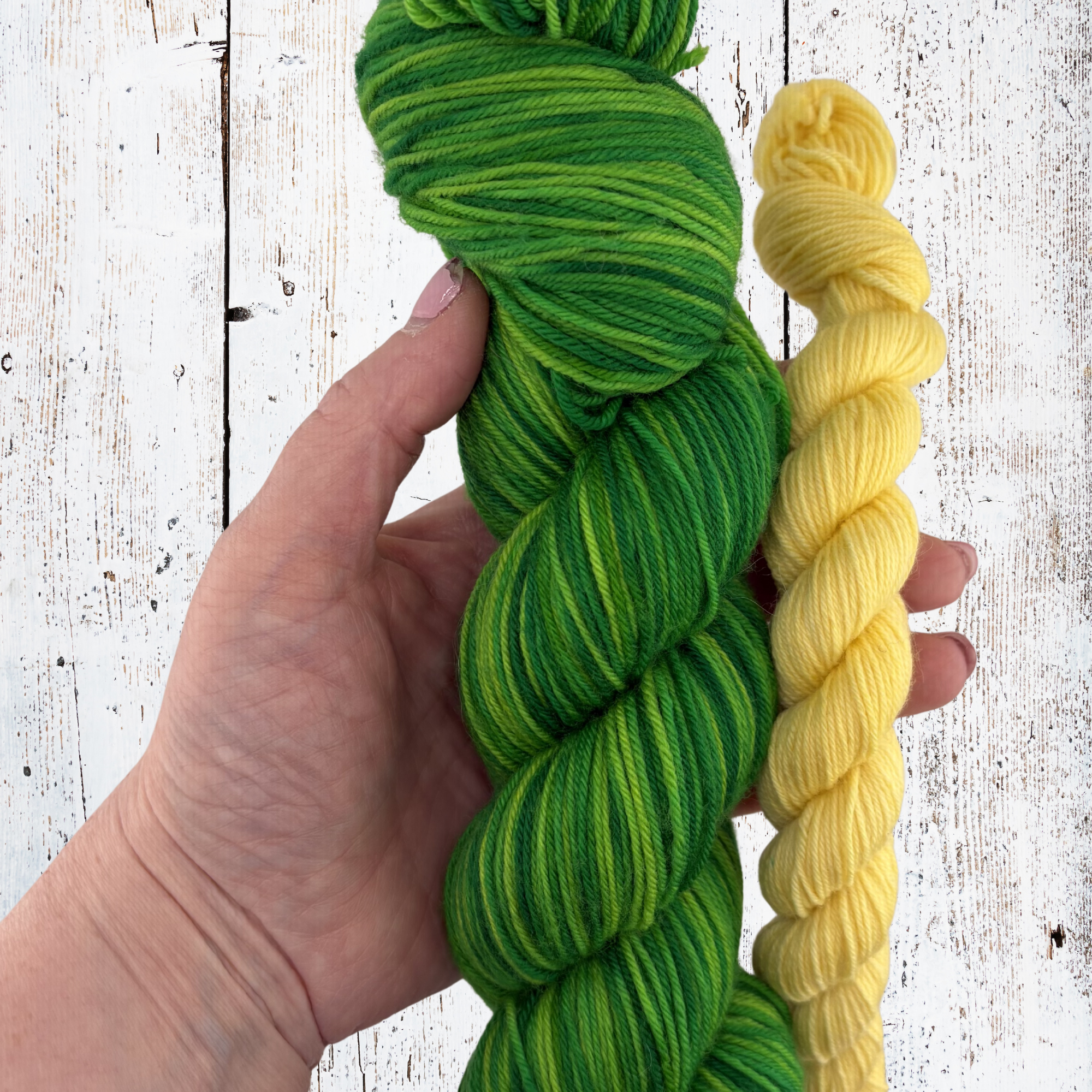 Green and yellow yarn skeins held against a white wooden background, highlighting tonal variation and smooth twist