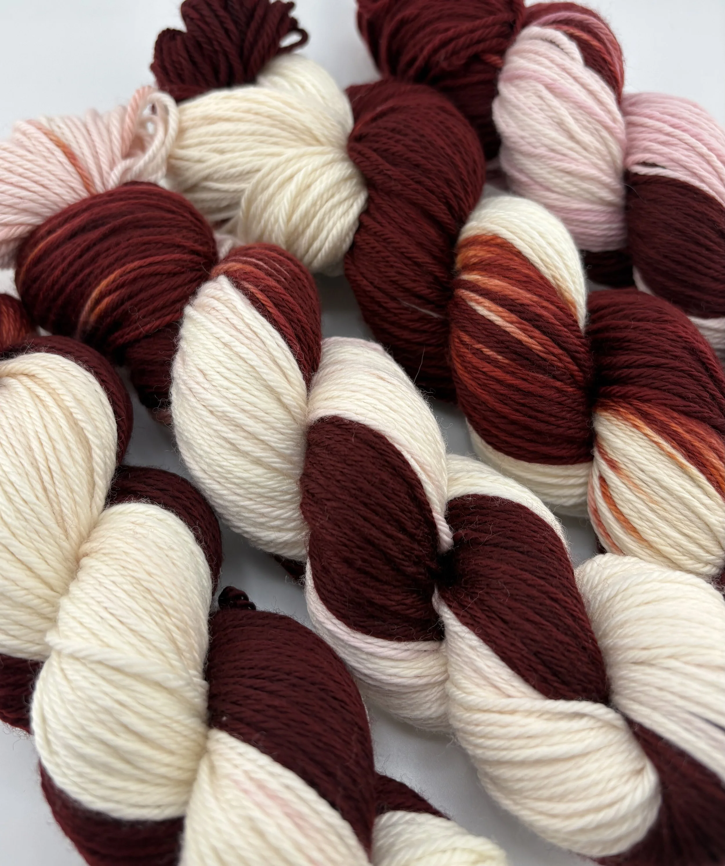 Hand‑dyed yarn skeins in cream and maroon and arranged together.”