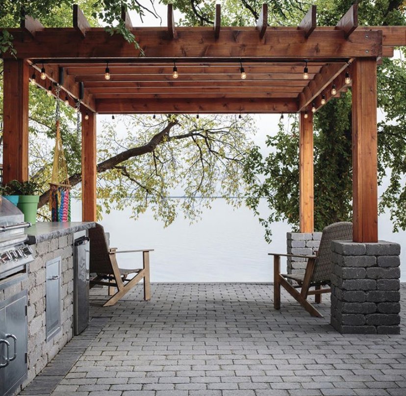 A cozy outdoor patio with a wooden pergola, string lights, two wooden chairs, a grill, and a view of water and trees in the background.