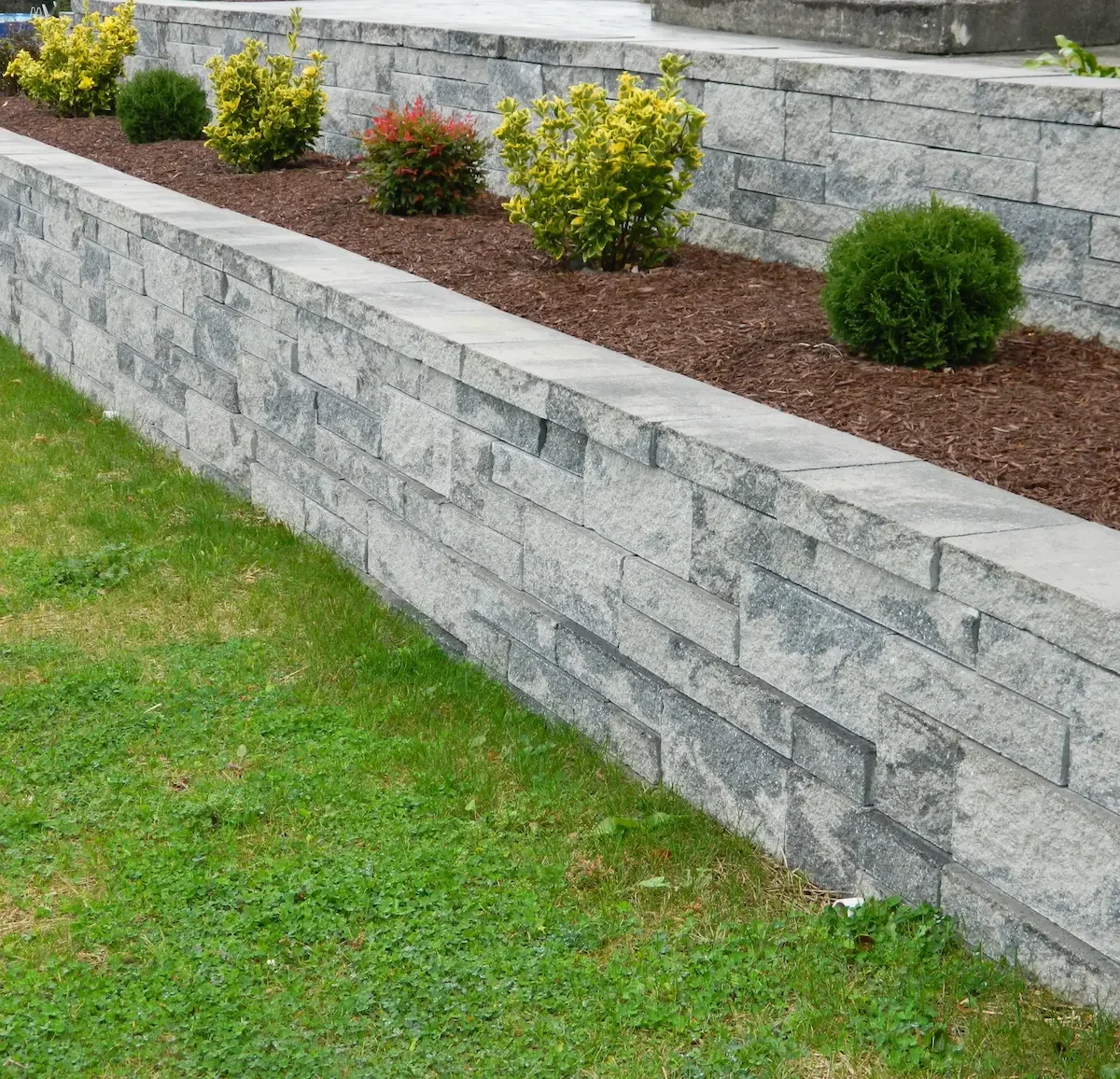 A stone retaining wall with a flower bed containing small bushes and mulch, bordered by a green grass lawn.