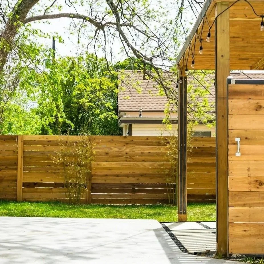 Backyard with wooden privacy fence, green grass, bright sunlight, and a small outdoor structure with string lights.