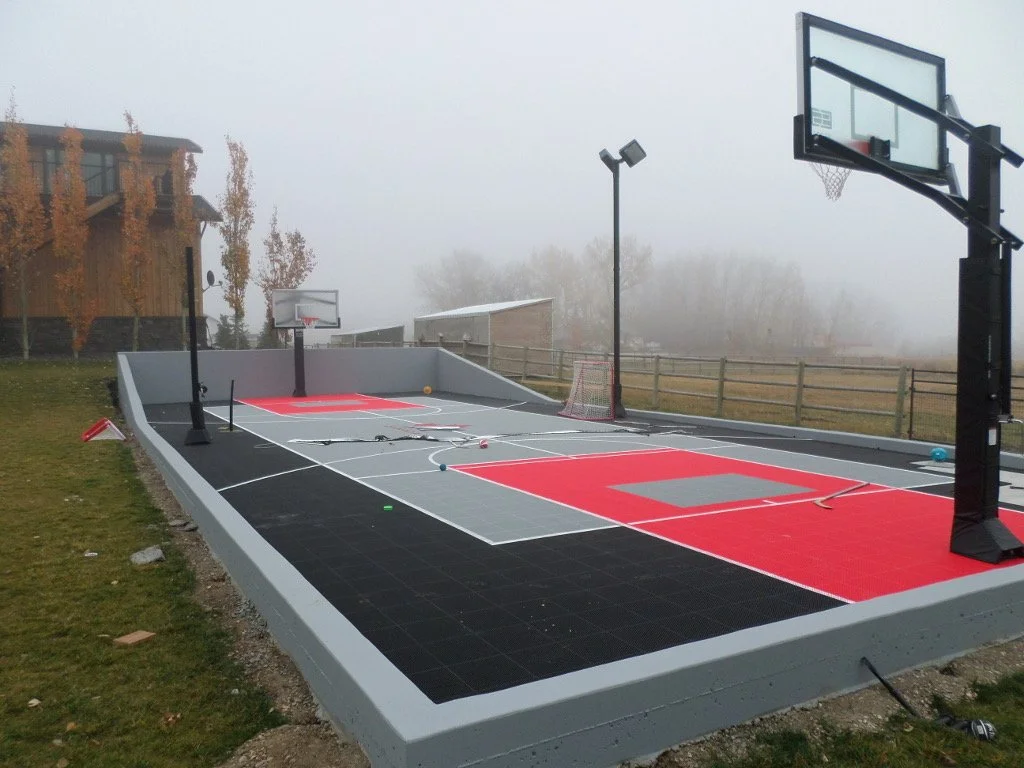 A small, outdoor basketball court with multiple hoops, surrounded by a foggyfield and a wooden fence, situated near a residential building with trees showing autumn foliage.