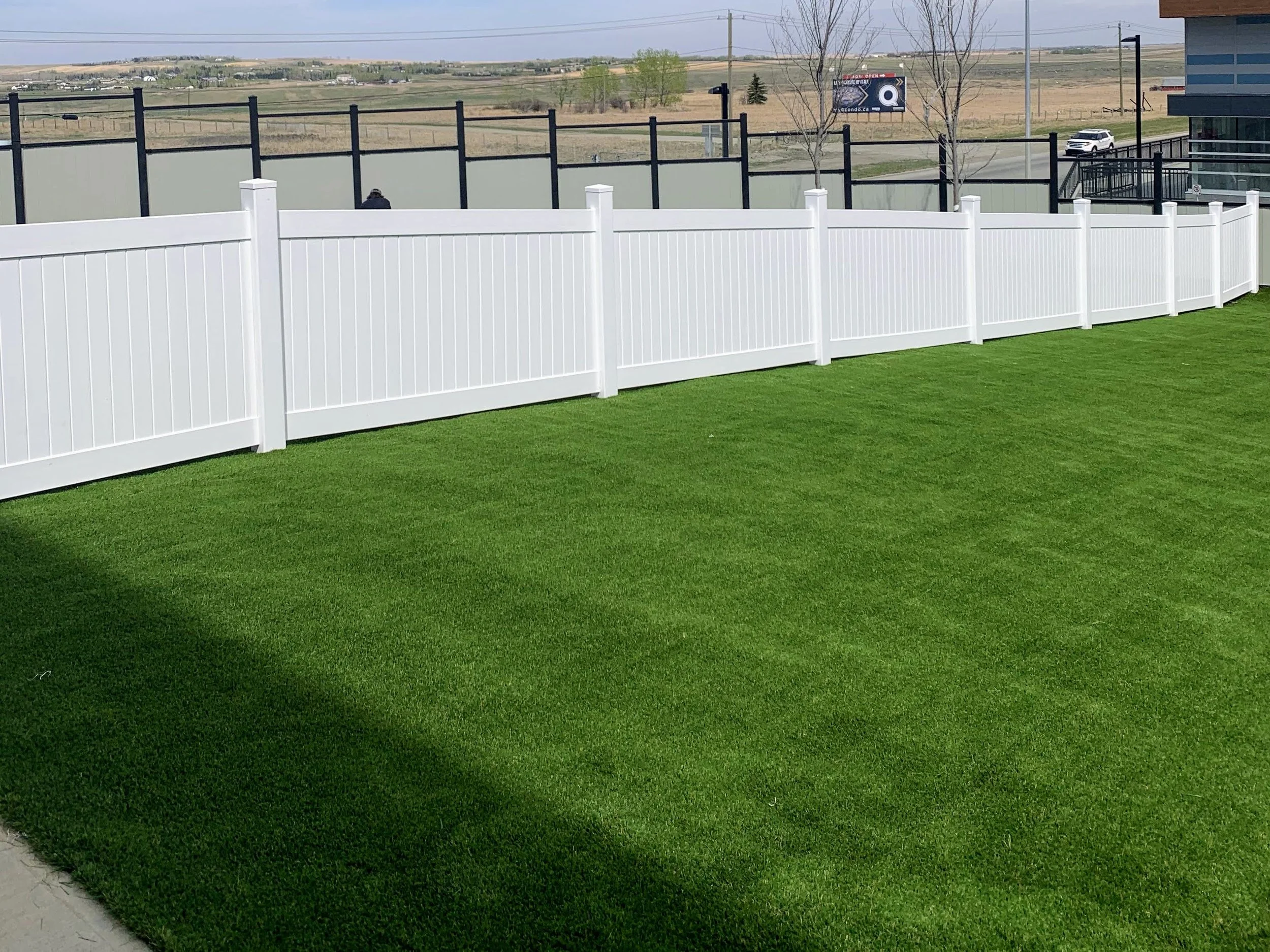 A backyard with bright green artificial grass, enclosed by a white vinyl fence, with another black fence and a parking lot visible in the background.