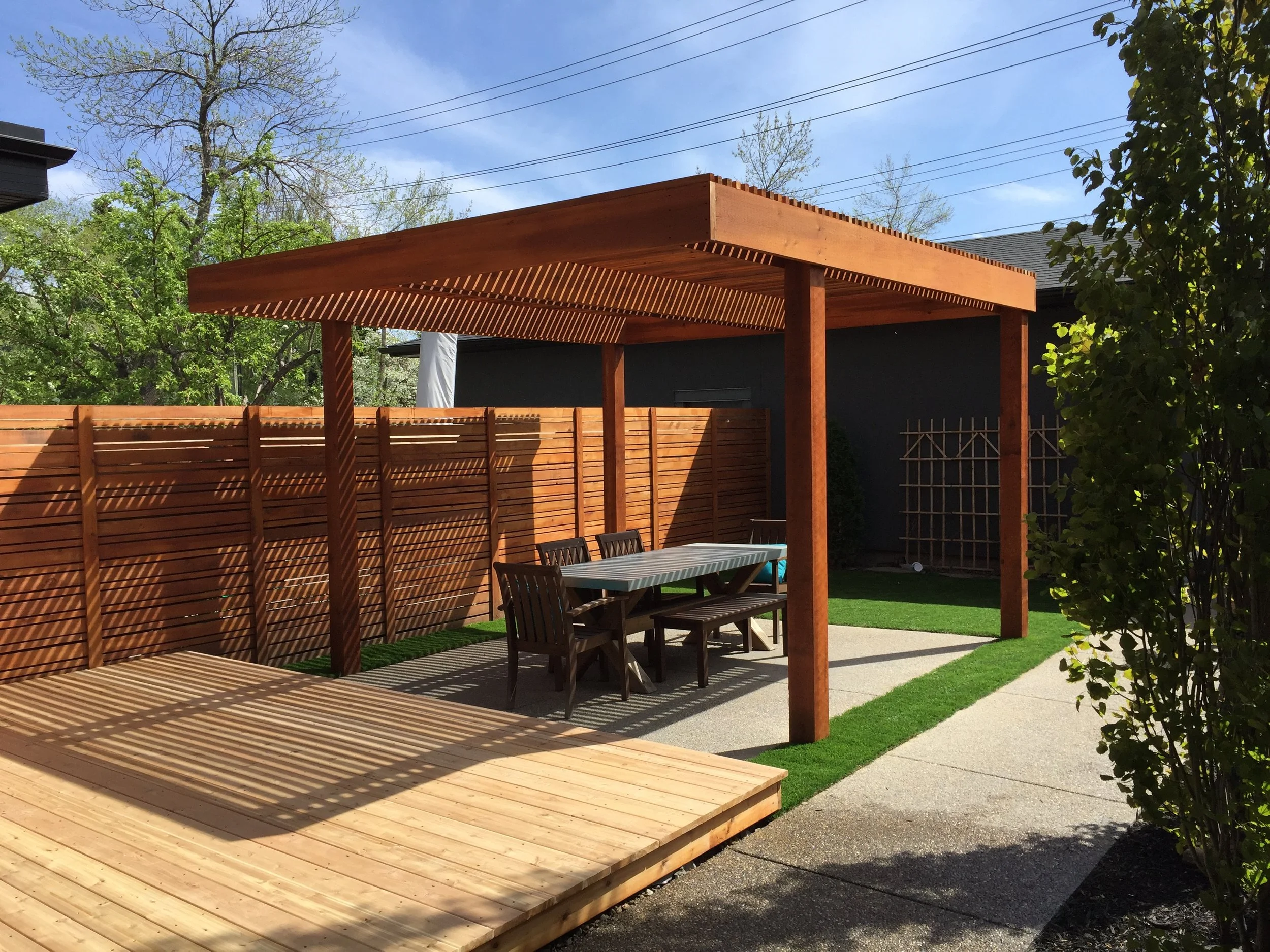 Backyard patio with a wooden pergola, outdoor dining table with chairs, wooden deck, and landscaped grass area.