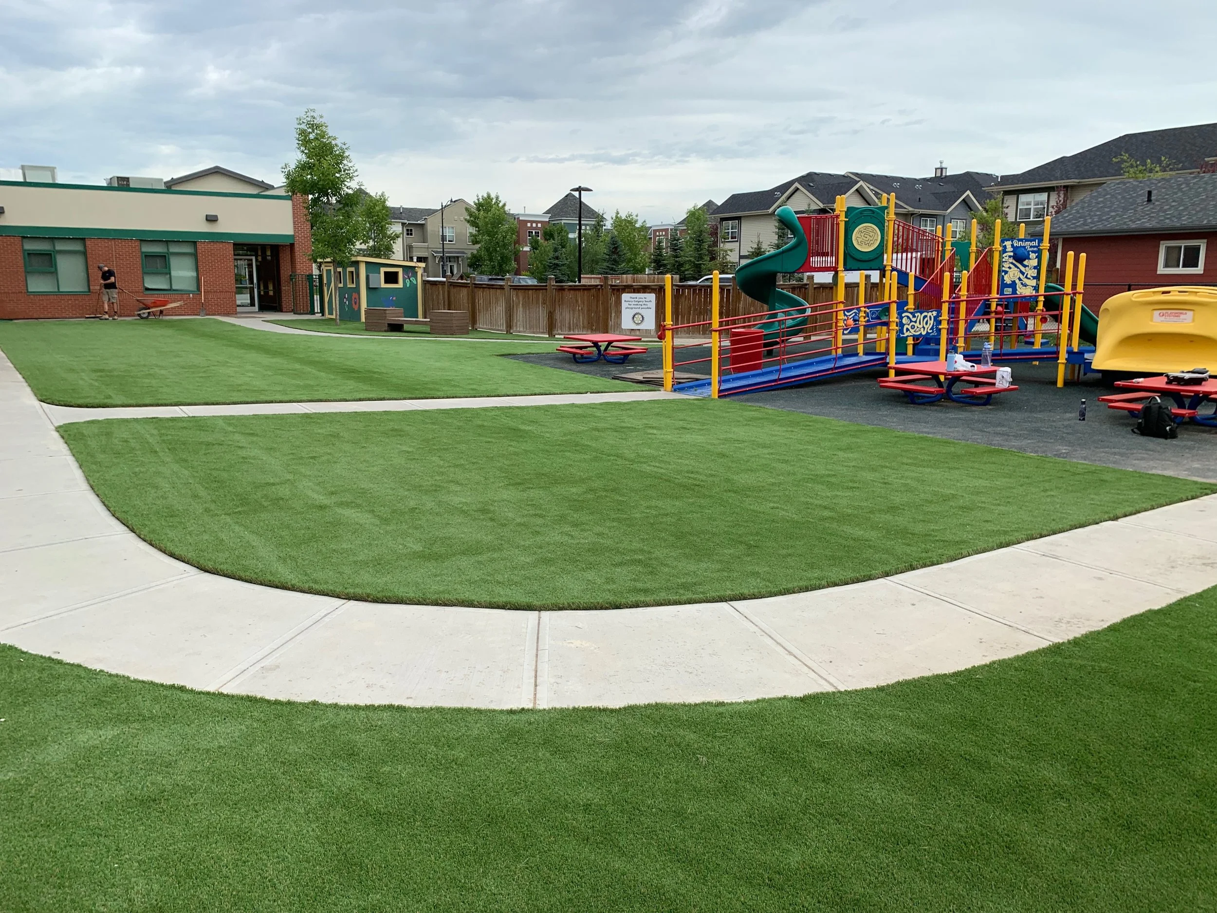 Playground with colorful equipment including slides and climbing structures, surrounded by grassy areas and concrete walkways in a residential neighborhood.