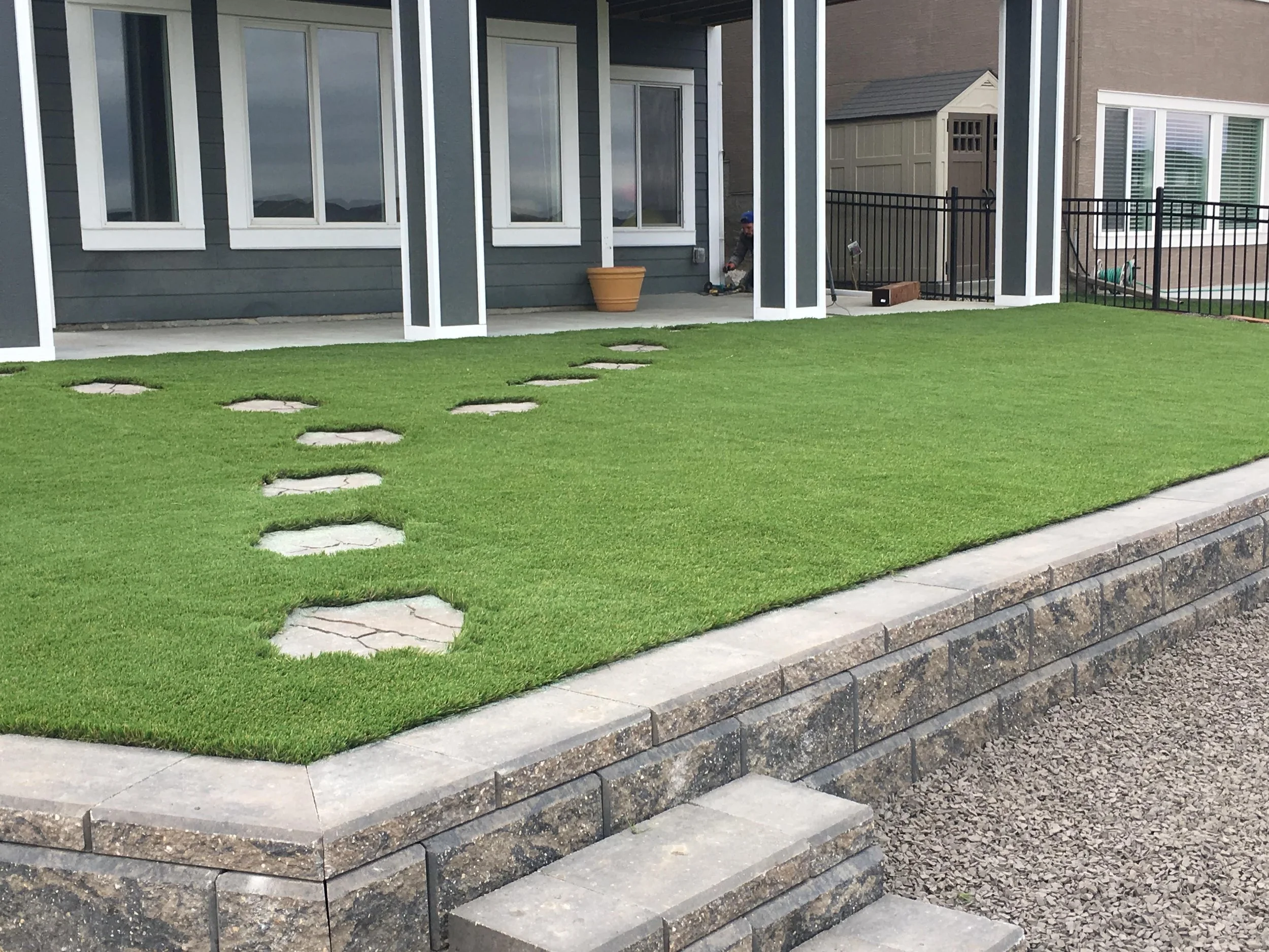 Backyard with green artificial turf, stone stepping stones in a curved line, stone steps leading up to a house, and a screened porch with a person working in the background.