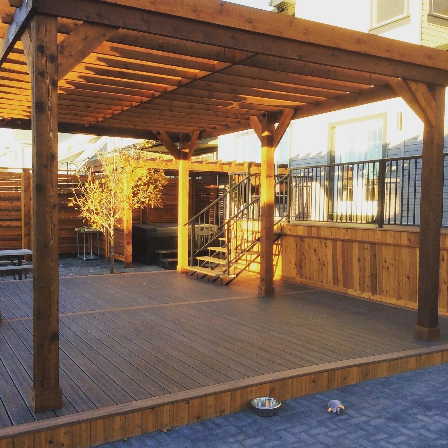 Empty wooden deck with a pergola, stairs leading up to a raised area, a small tree, a metal railing, a bowl, and a small plush toy on the ground.