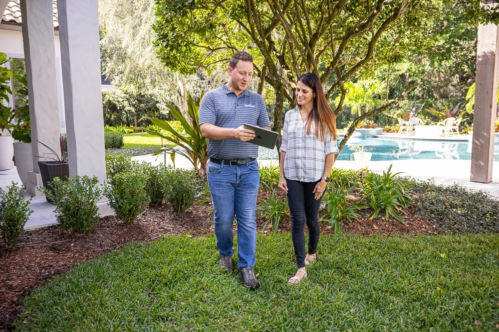 Man showing tablet to woman in a backyard garden with trees and pool in the background.
