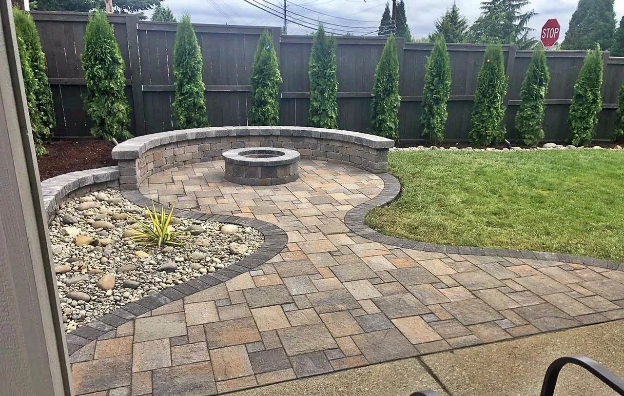 A backyard patio with brick pavers, a stone fire pit, a curved low brick wall, a grassy area, and a row of tall, narrow evergreen trees along a wooden fence. A stop sign is visible in the background.