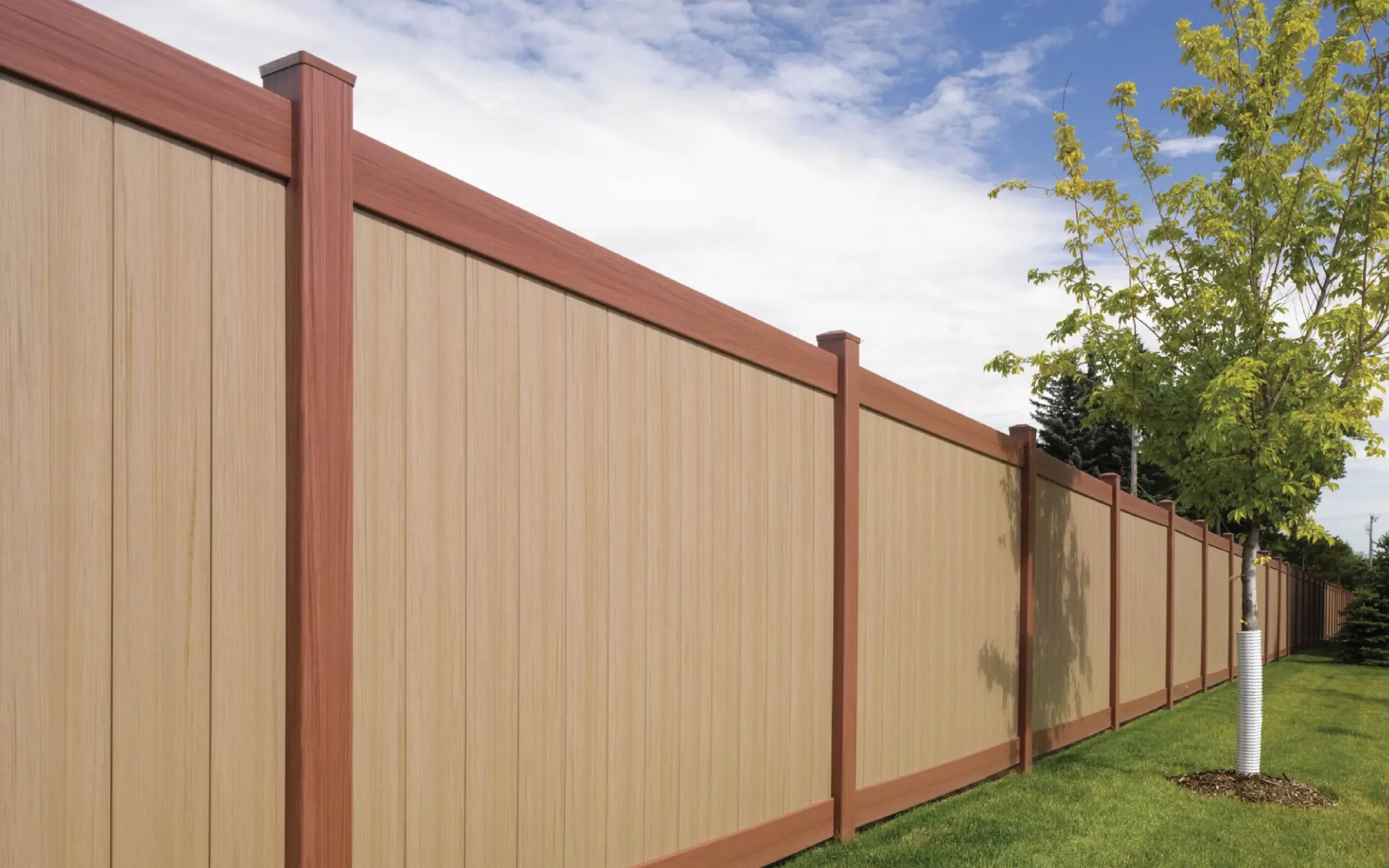 A beige and brown wooden privacy fence runs along a green lawn with a small tree in front of it. The sky is partly cloudy.