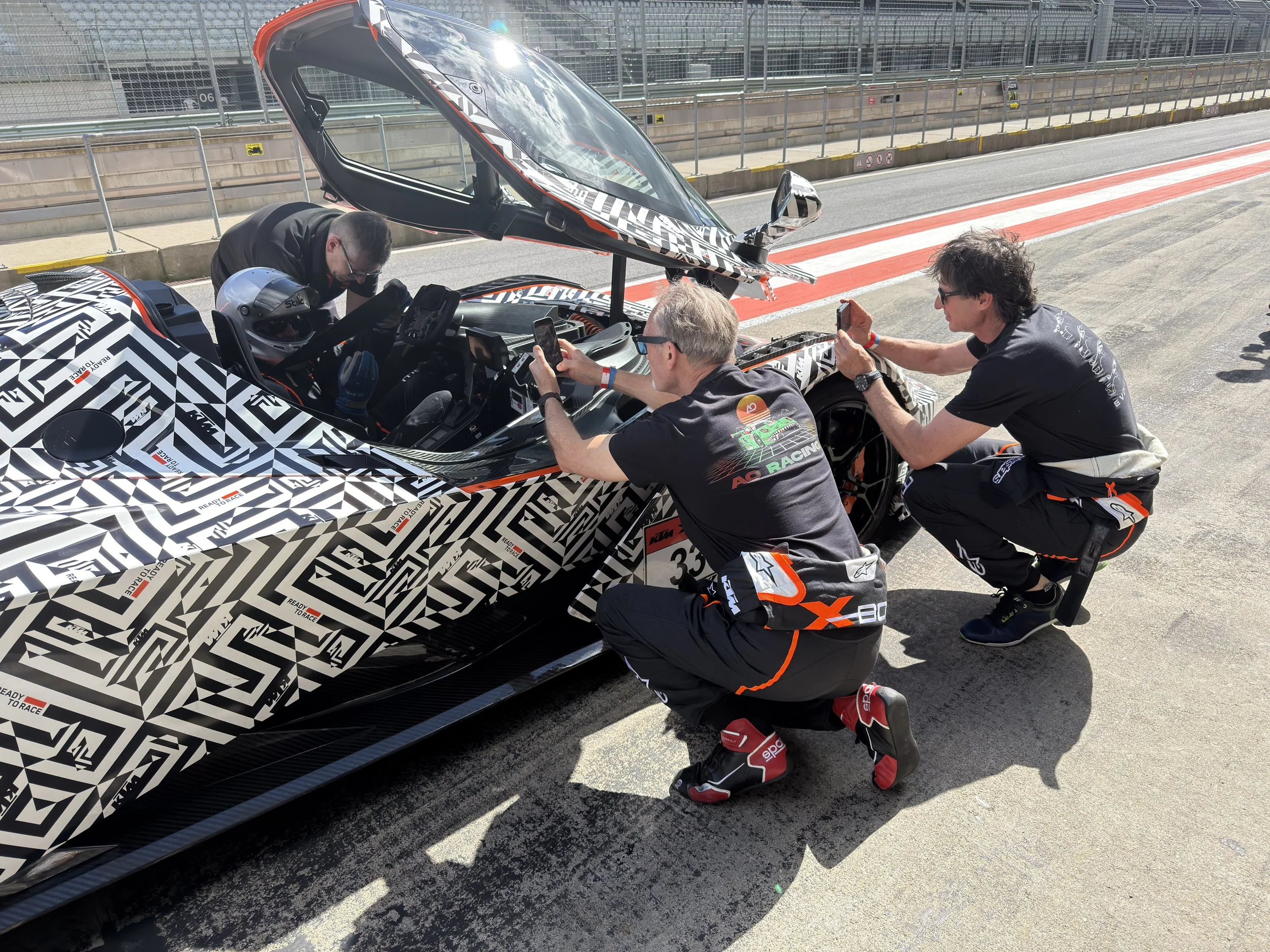 Steven Madge in a KTM XBOW wearing in racing suits and casual clothes working on a black and white patterned racing car on a race track. One man is inside the car, two men are kneeling outside, taking photos and inspecting.