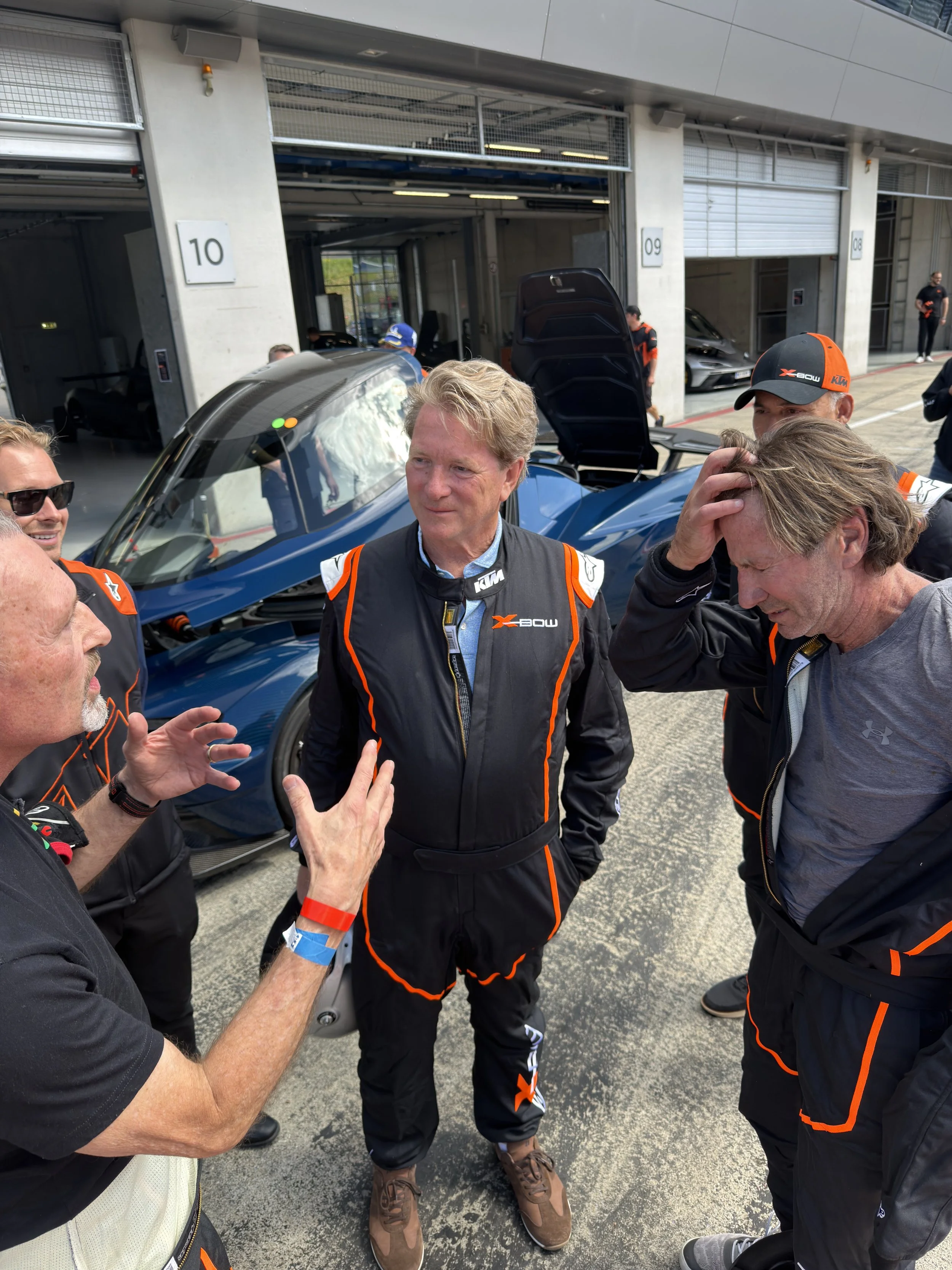 Group of people at a racing event, standing in front of a blue racing car in a garage area, talking and gesturing.