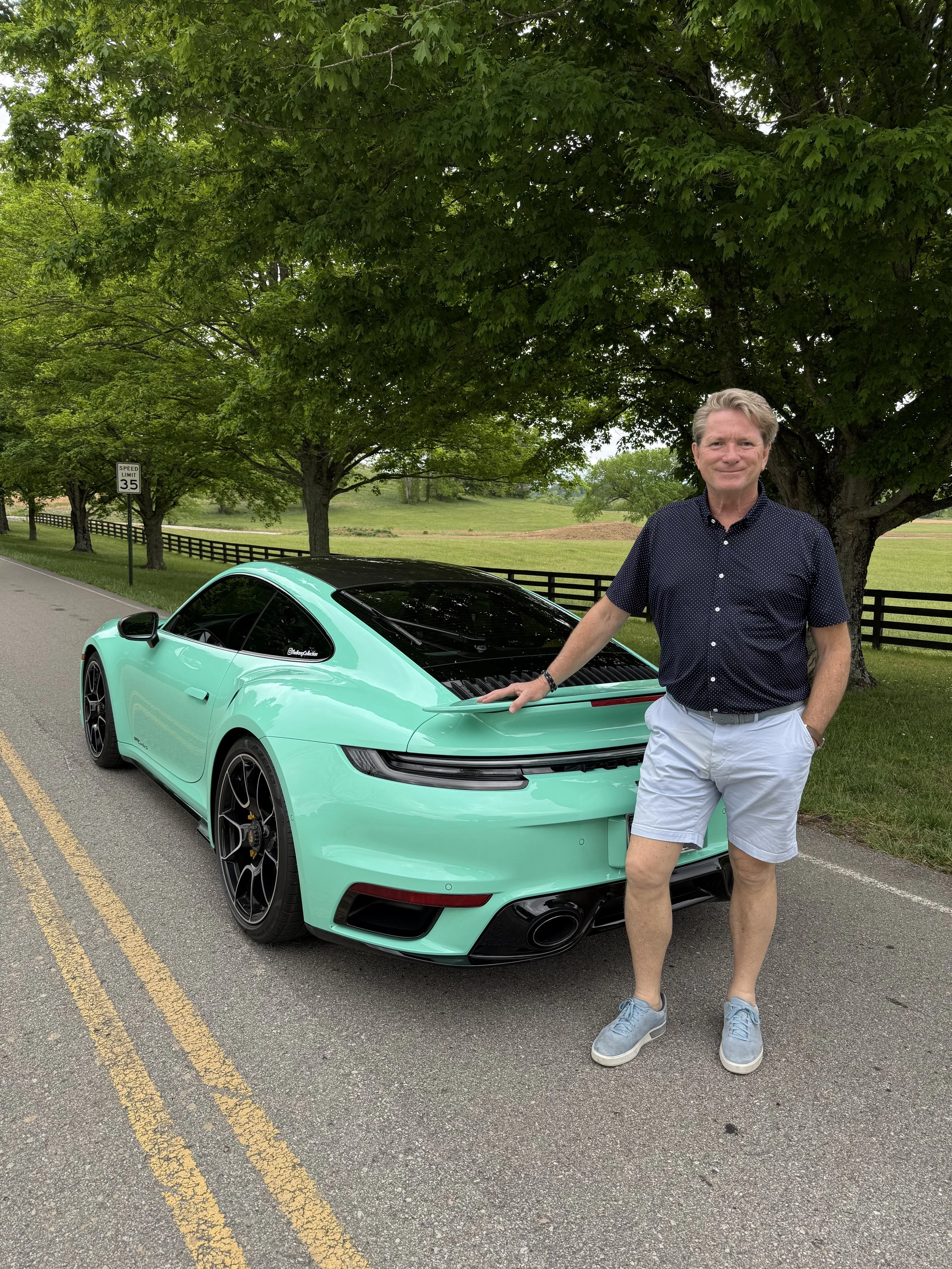 Steven Madge standing next to a mint green porsche turbo s with trees and a green field in the background. The man is wearing a dark blue, short-sleeved, button-up shirt, white shorts, and gray sneakers, and is smiling at the camera.