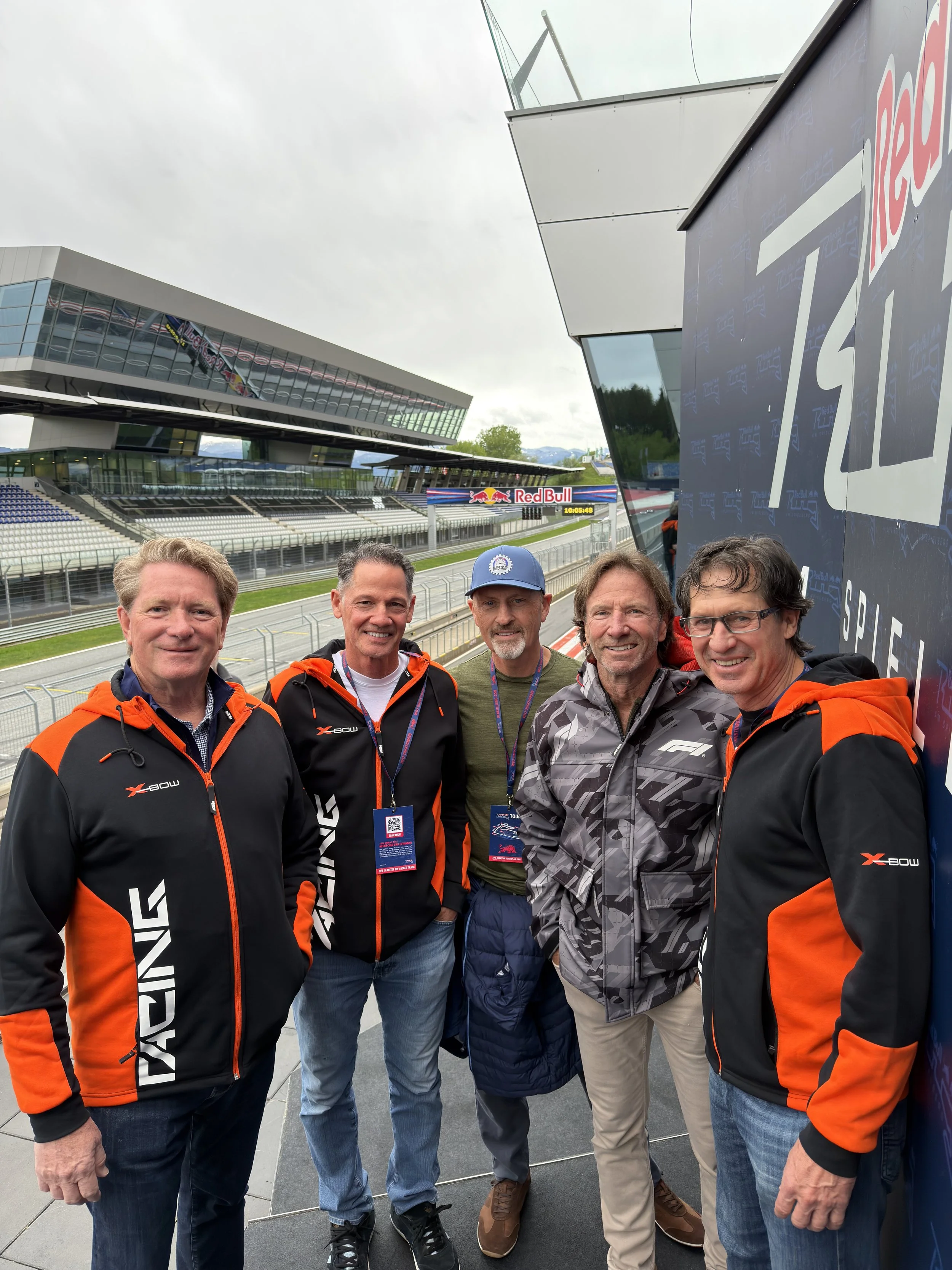 Five men standing together outdoors at a racing event near a racetrack and grandstand, smiling at the camera.