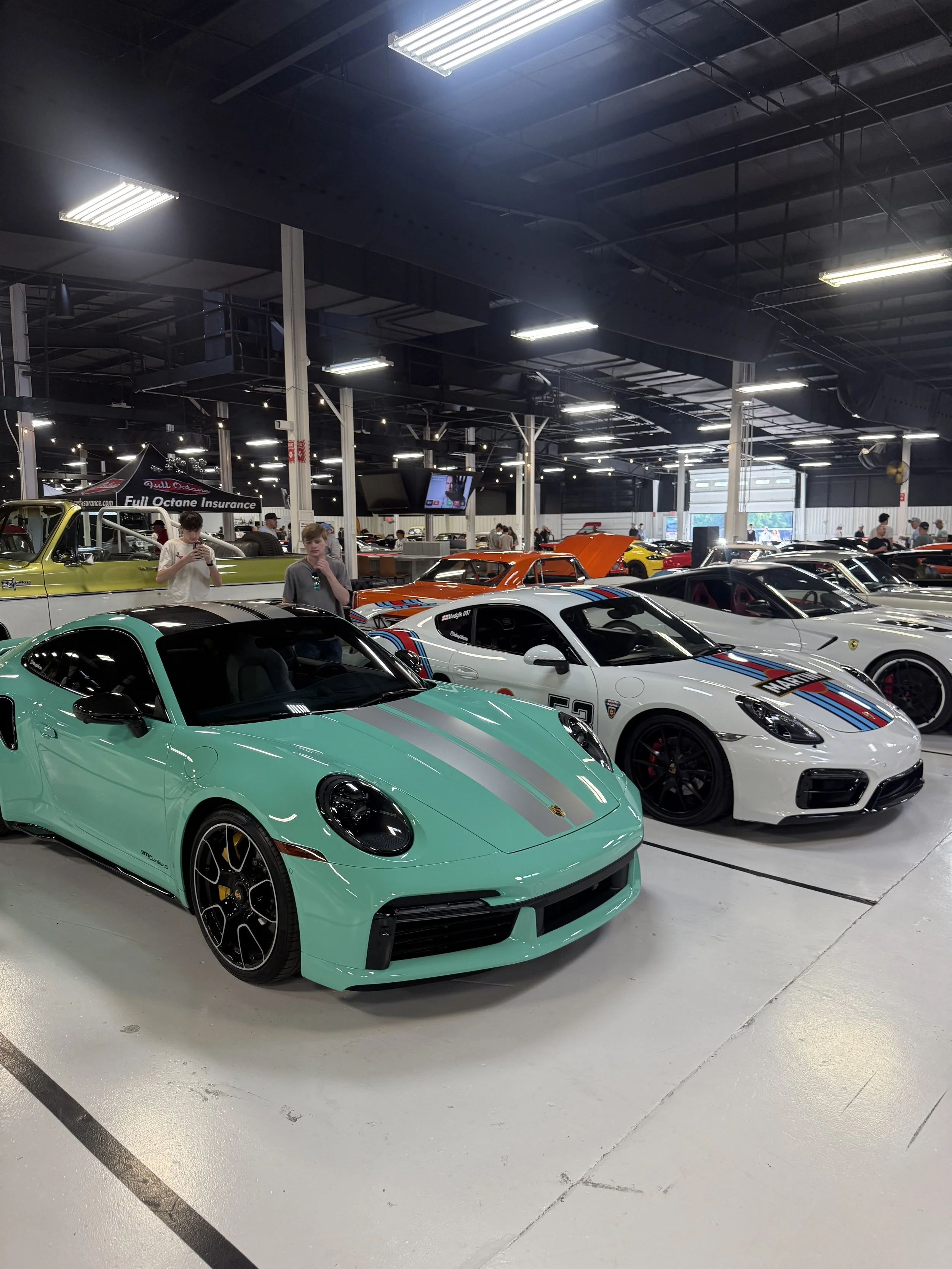 Multiple luxury sports cars on display inside the 111 Motor Cars showroom. Prominent porsche mint green and white Porsche with a racing stripe, other colorful sports cars are visible in the background.