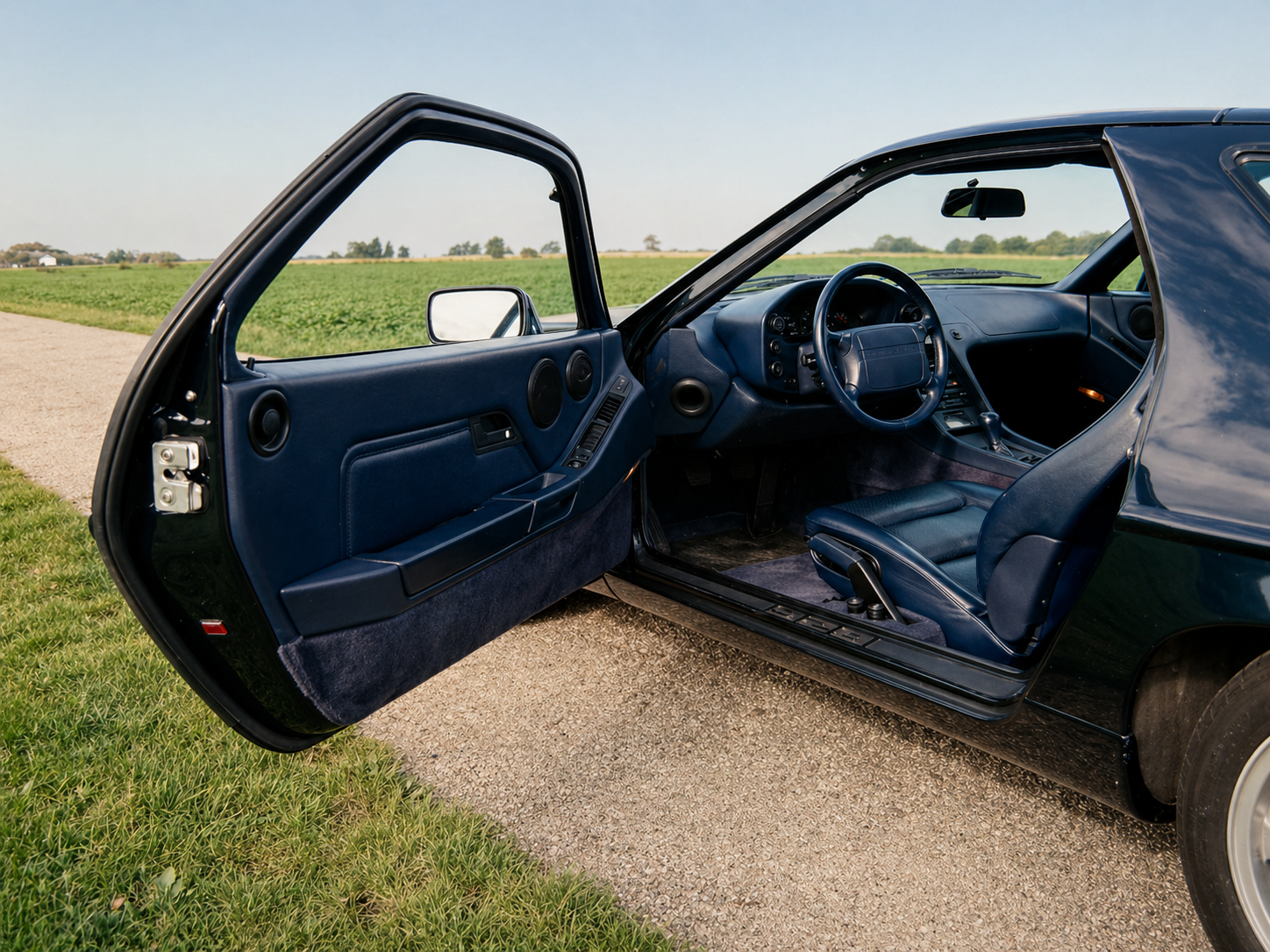 Porsche 928 GTS Open door revealing interior of a black sports car with blue leather seats, dashboard, steering wheel, and control panel, parked beside a grassy field and gravel road.