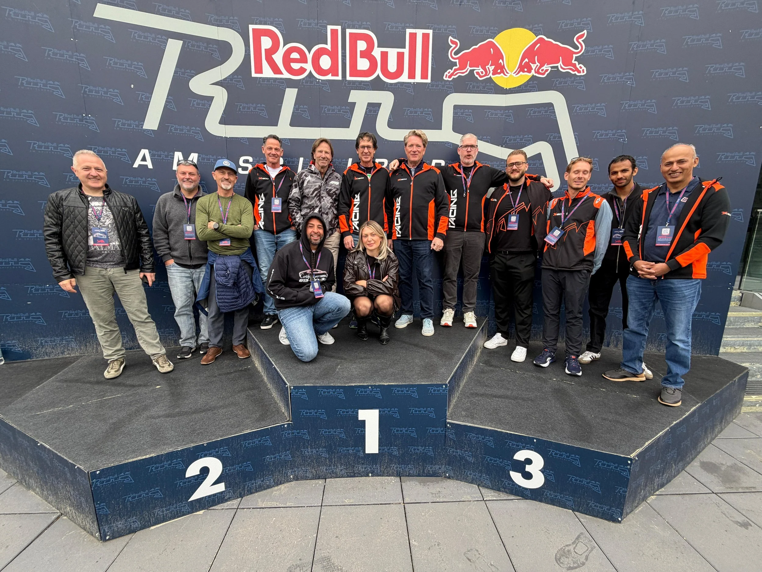 Group of people standing on a winner's podium in front of a large Red Bull logo and racing track graphic backdrop, smiling for a photo.