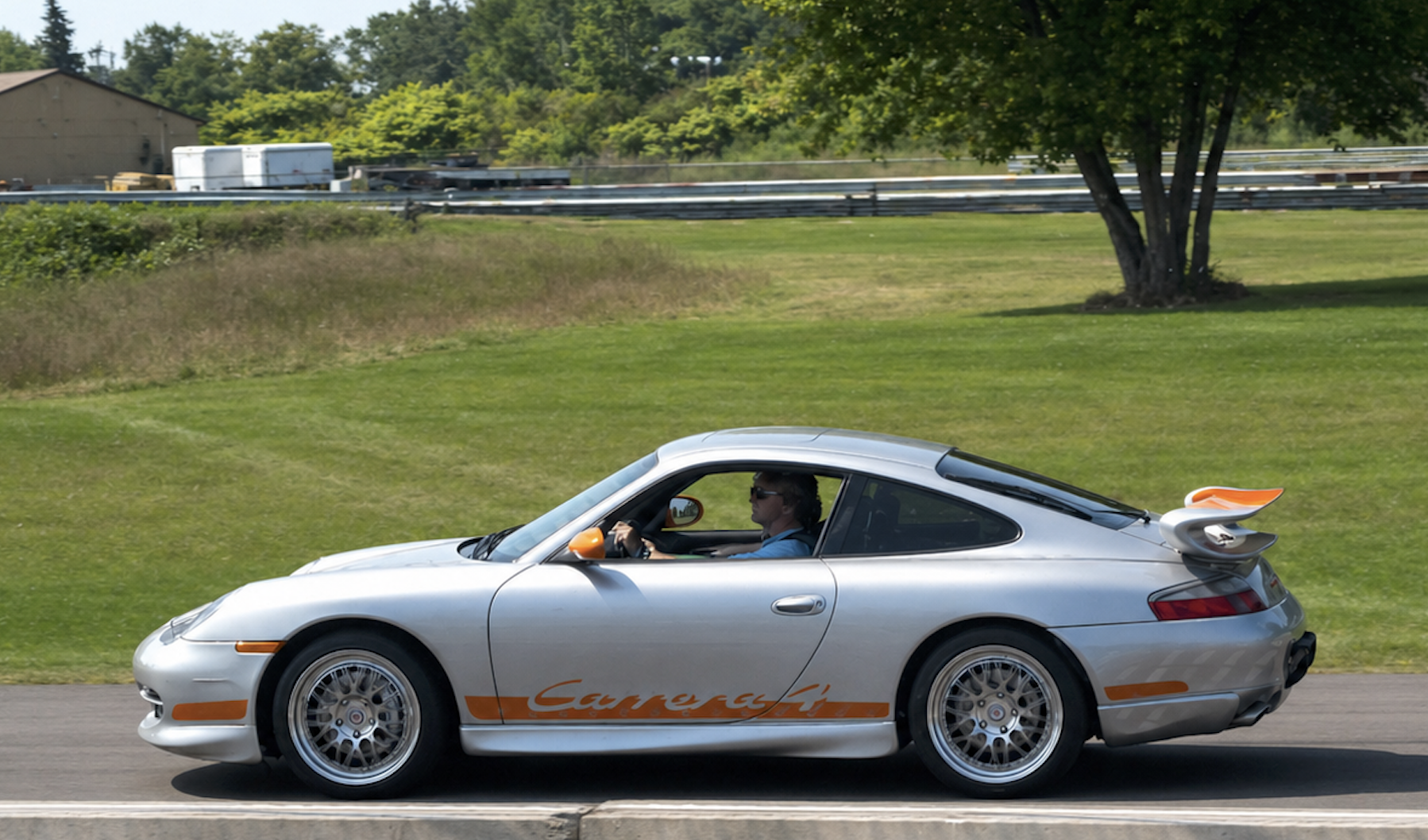 A silver Porsche Carrera with orange accents driving on a road next to a grassy area with trees, a building, and a highway in the background. The driver is wearing sunglasses.