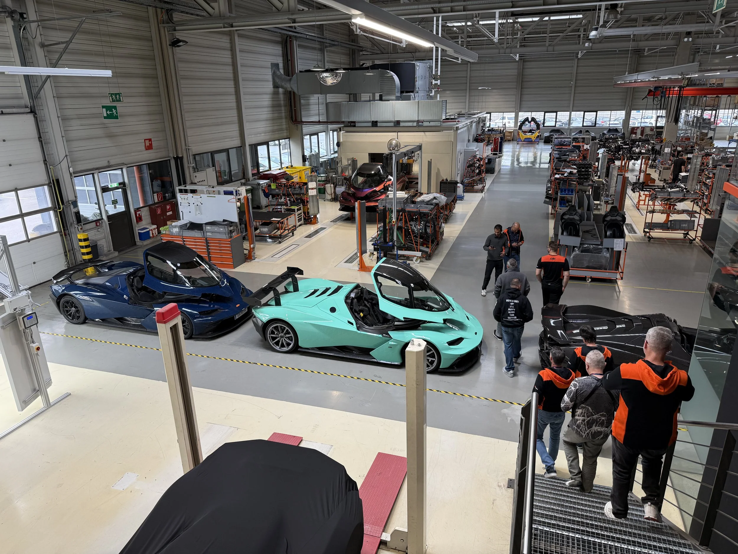 Multiple KTM X-Bow sports cars, including a blue, green, and black vehicle, parked inside the KTM factory in Austria.
