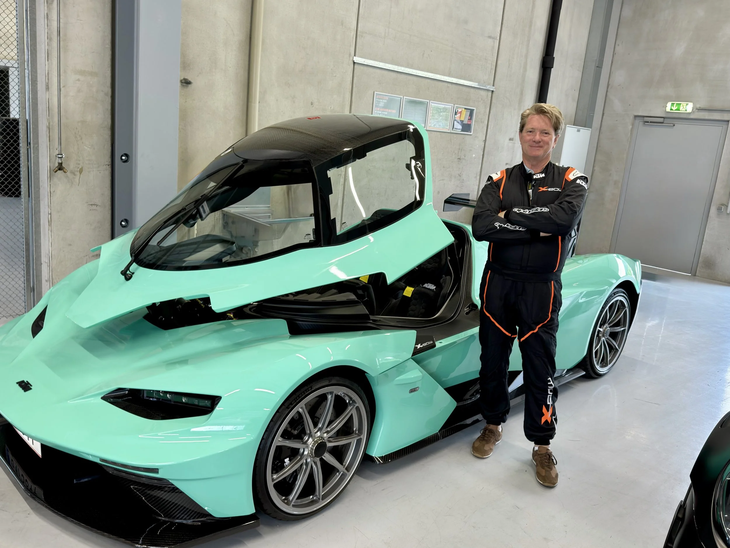 Steven Madge dressed in a racing suit standing next to a Mint green KTM X-BOW sports car at the Red Bull Race track in Austria.