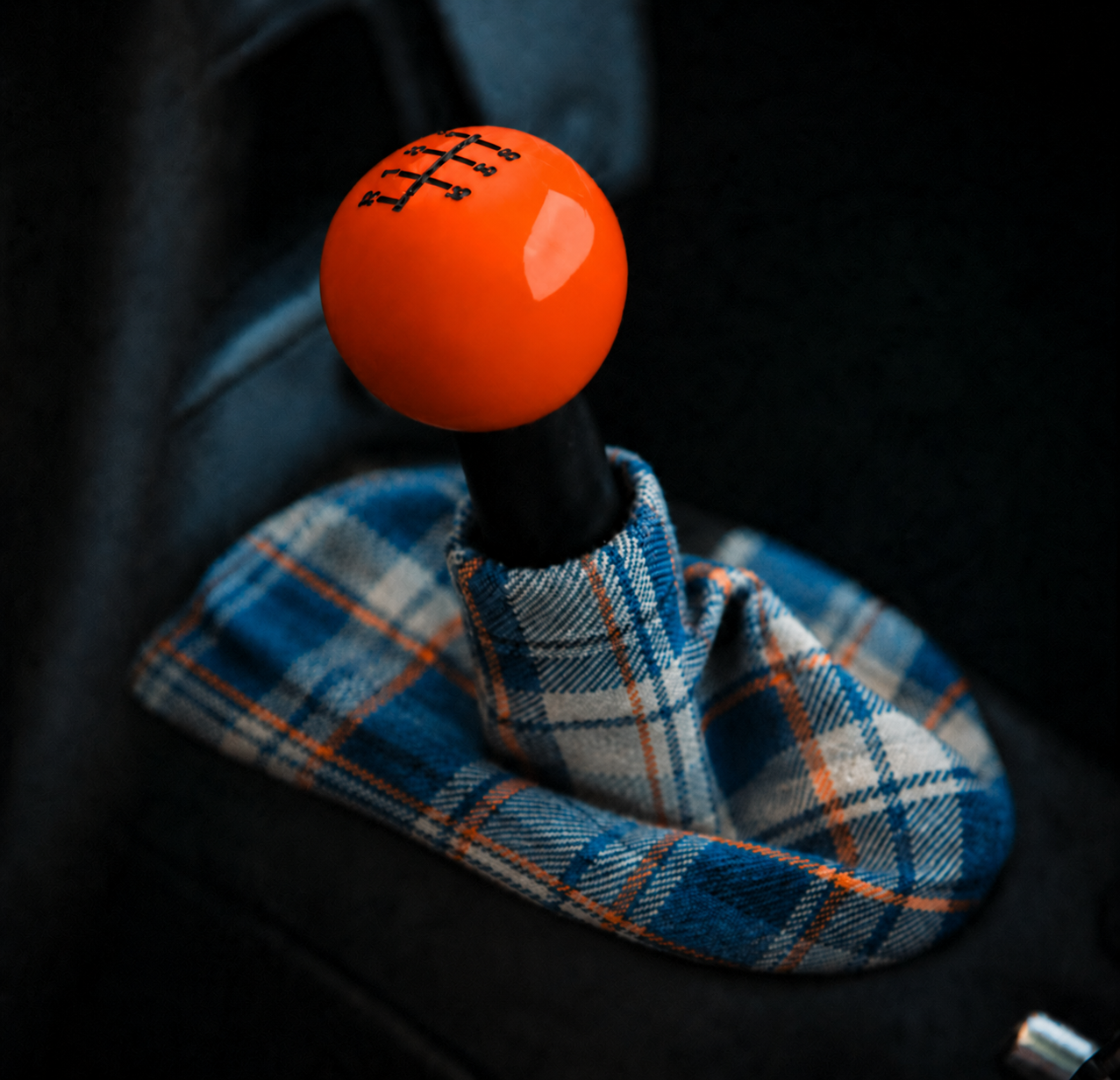 A gear shift knob with an orange soccer ball design on top, placed in a vehicle's interior with a blue, white, and orange plaid fabric around the base.