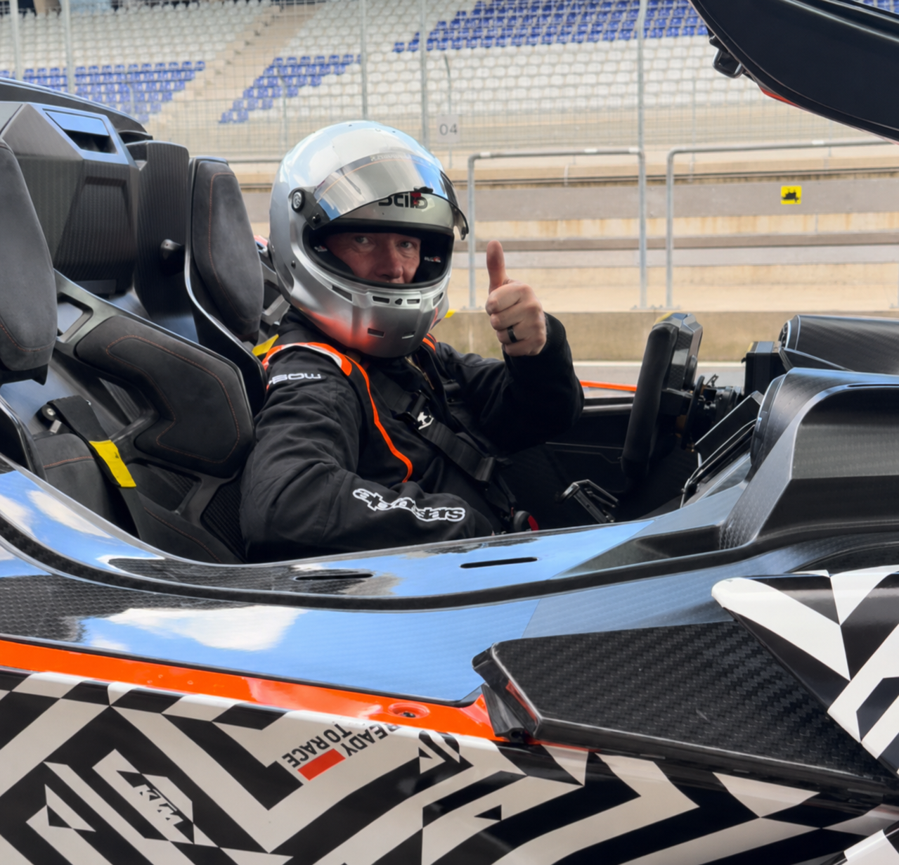 Steven Madge  sitting in a high-performance KTM X-BOW race car, wearing a helmet and black racing suit, giving a thumbs-up gesture on a race track.