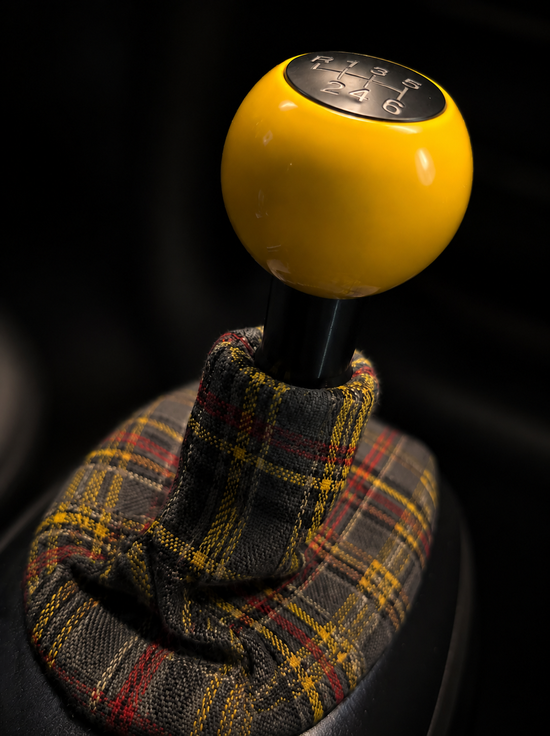 Close-up of a yellow car gear shift knob with black top showing a shift pattern, held by a hand in a plaid glove.