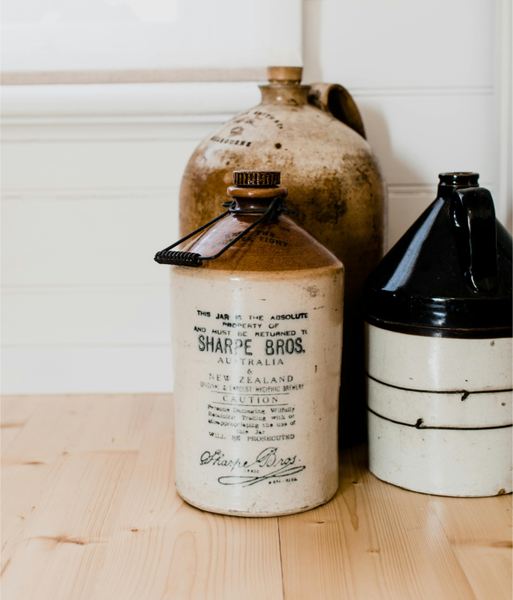 Antique ceramic bottles and jars on a wooden floor against a white wall, with one labeled 'Sharpe Bros' from Australia and New Zealand.