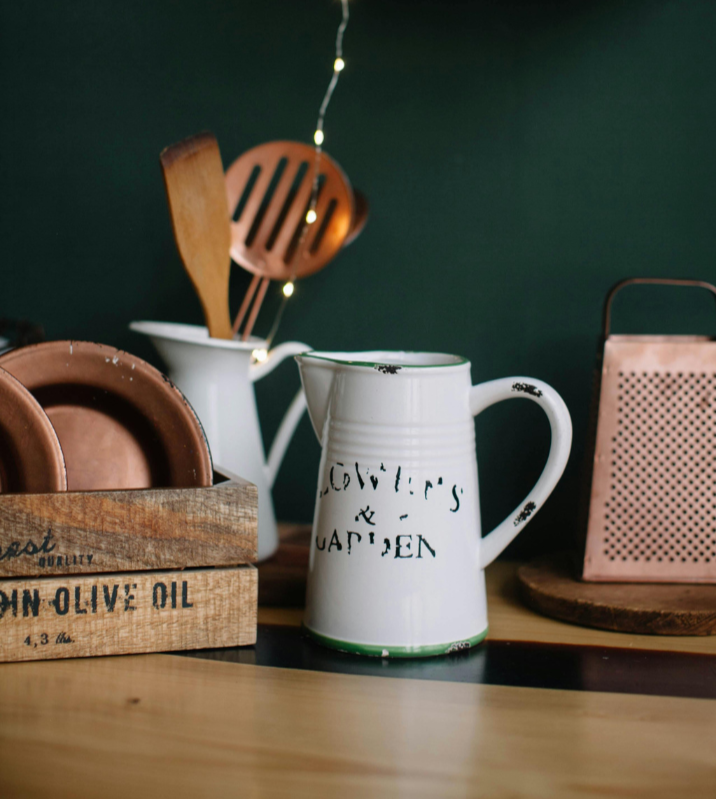 Kitchen countertop with a white mug labeled "Growing & Cappeden," a wooden box with copper-colored plates, a white pitcher with kitchen utensils, a pink metal container, and a wooden cutting board on a dark green wall background.