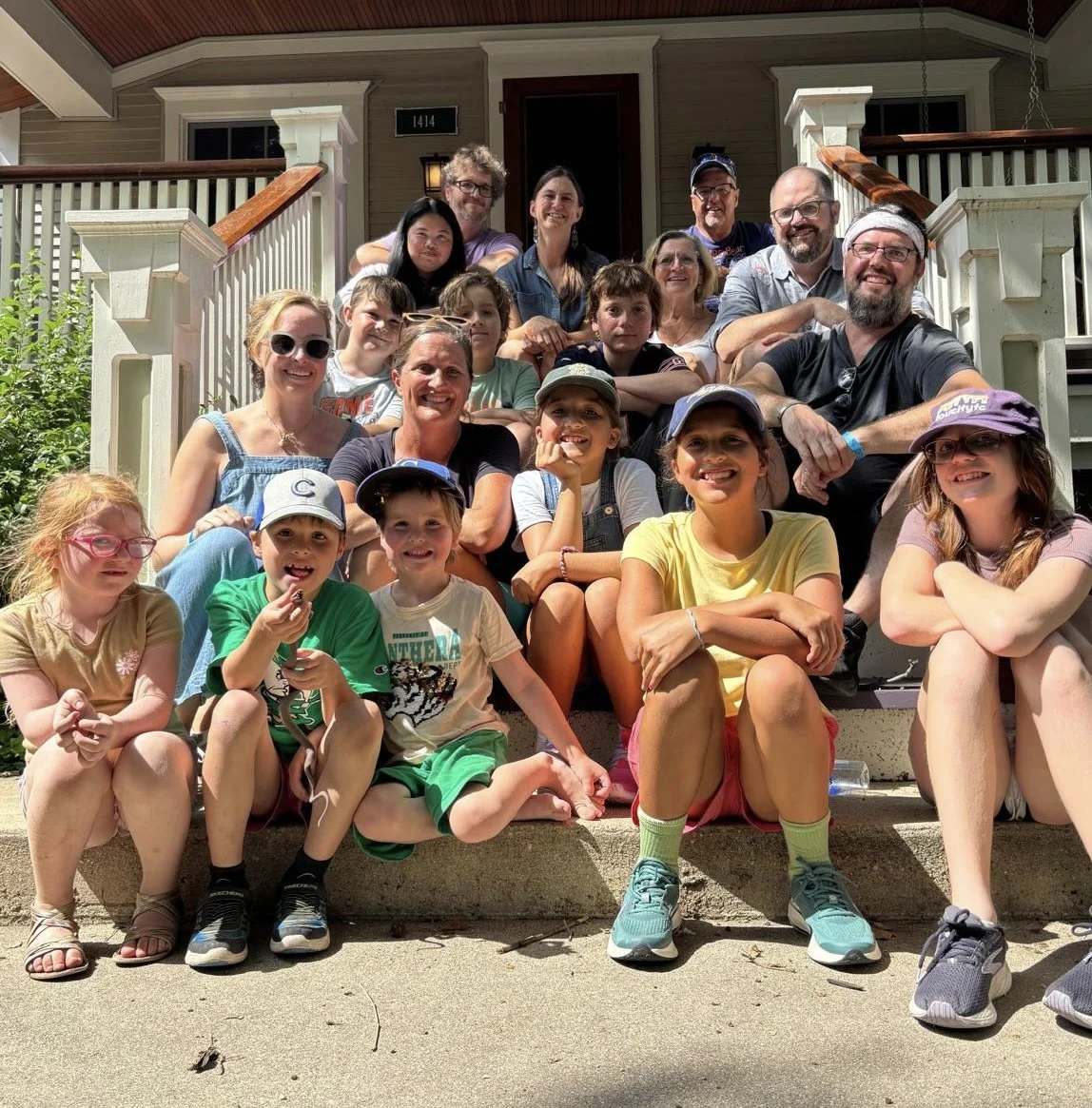Group of children and adults sitting on front steps of a house, smiling for a photo on a sunny day.