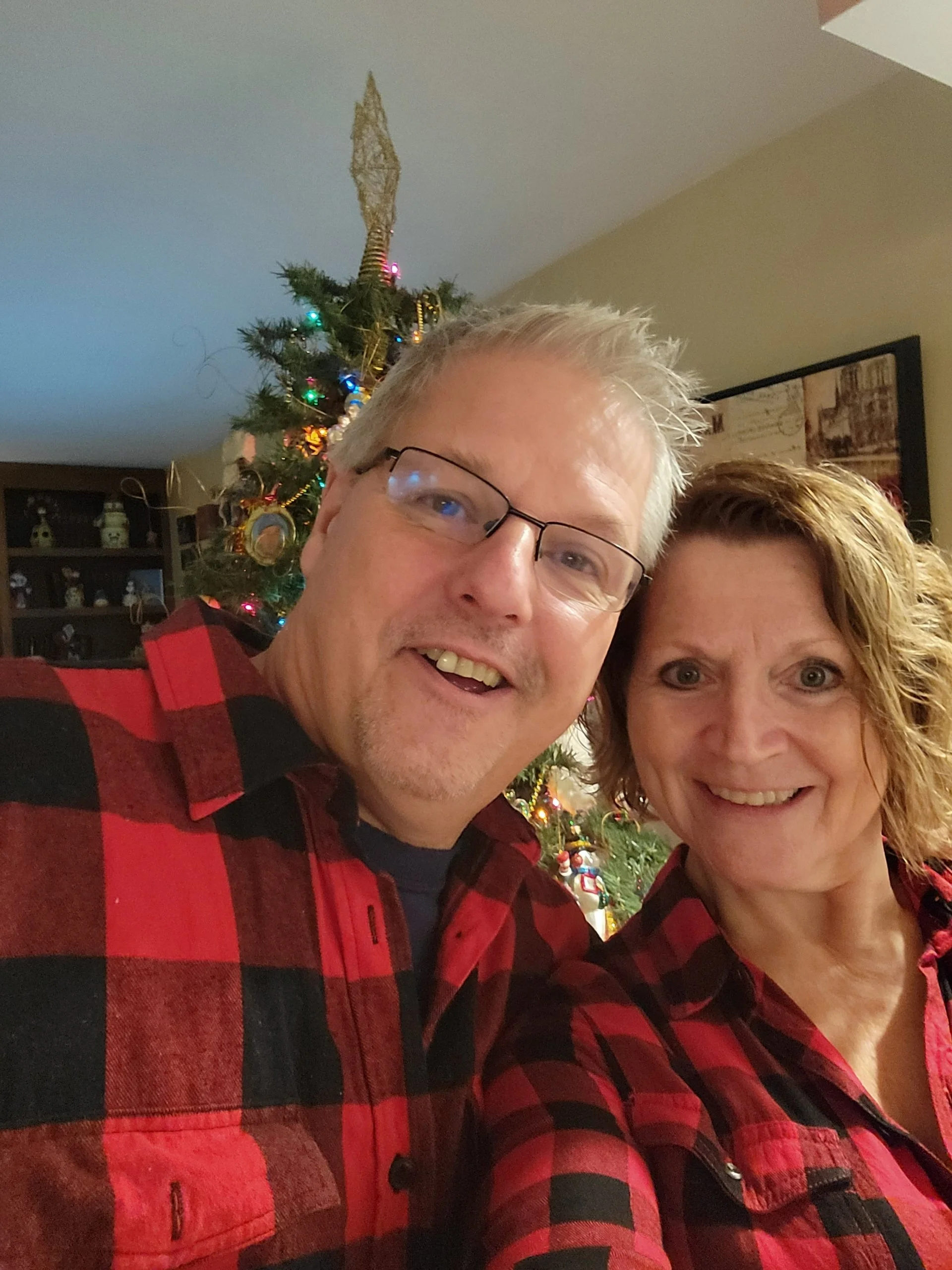 A smiling man and woman wearing matching red and black plaid shirts taking a selfie in front of a decorated Christmas tree.