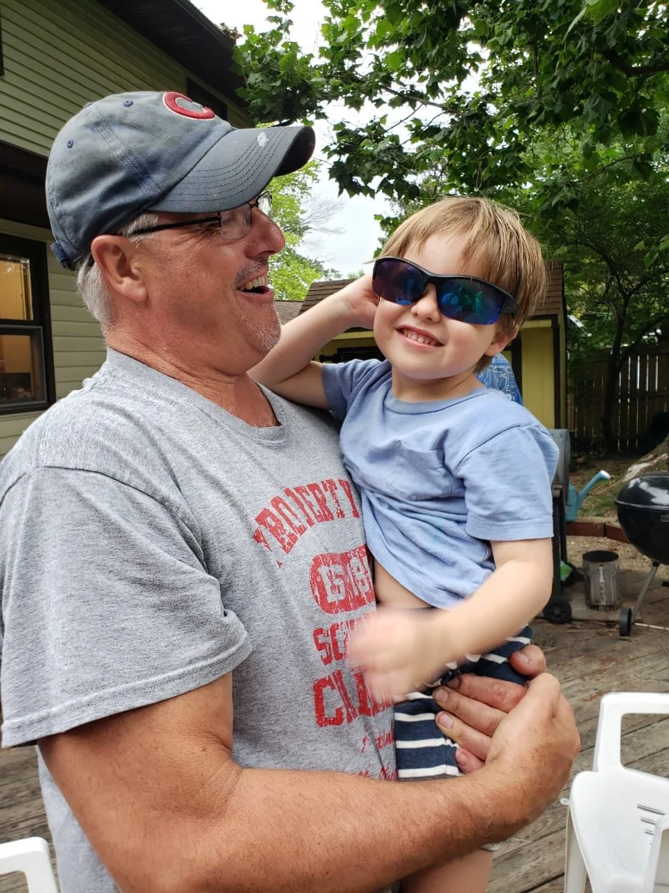 An older man with glasses and a baseball cap holding a young boy with sunglasses and a blue shirt outside on a wooden deck surrounded by trees and backyard items.