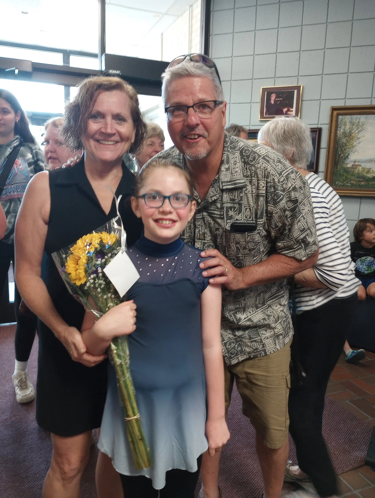 A young girl holding a bouquet of yellow flowers, standing with a black sleeveless dress, glasses, and smiling, flanked by a woman and a man in an indoor setting with other people and framed artwork on the wall.