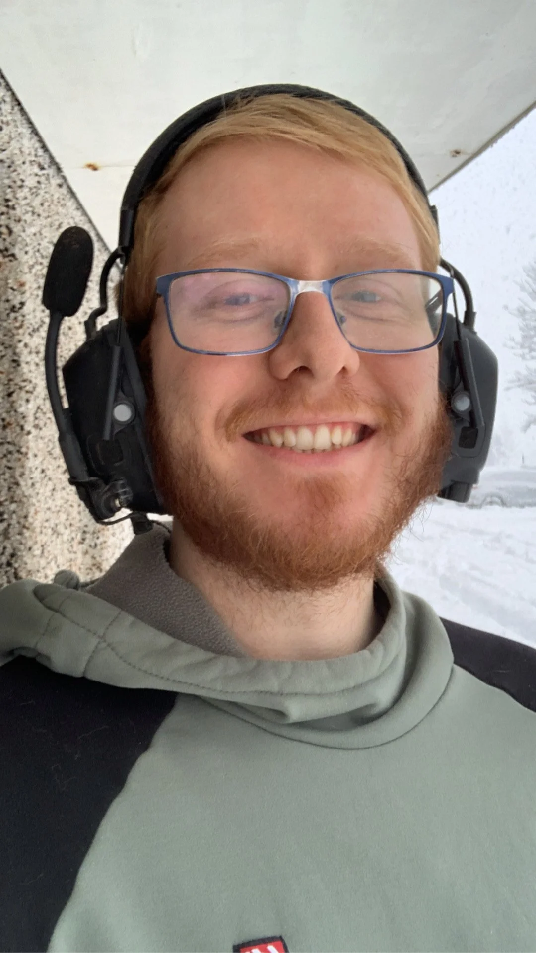 A smiling man wearing glasses, a headset, and a gray hoodie outdoors with snow and trees in the background.
